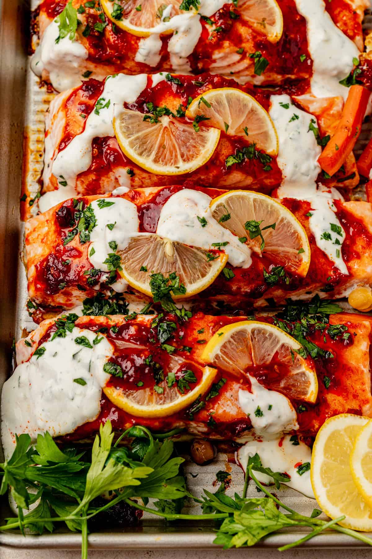 Baked harissa salmon fillets topped with lemon slices, white sauce, and herbs, arranged in a baking tray with fresh parsley and lemon wedges on the side.