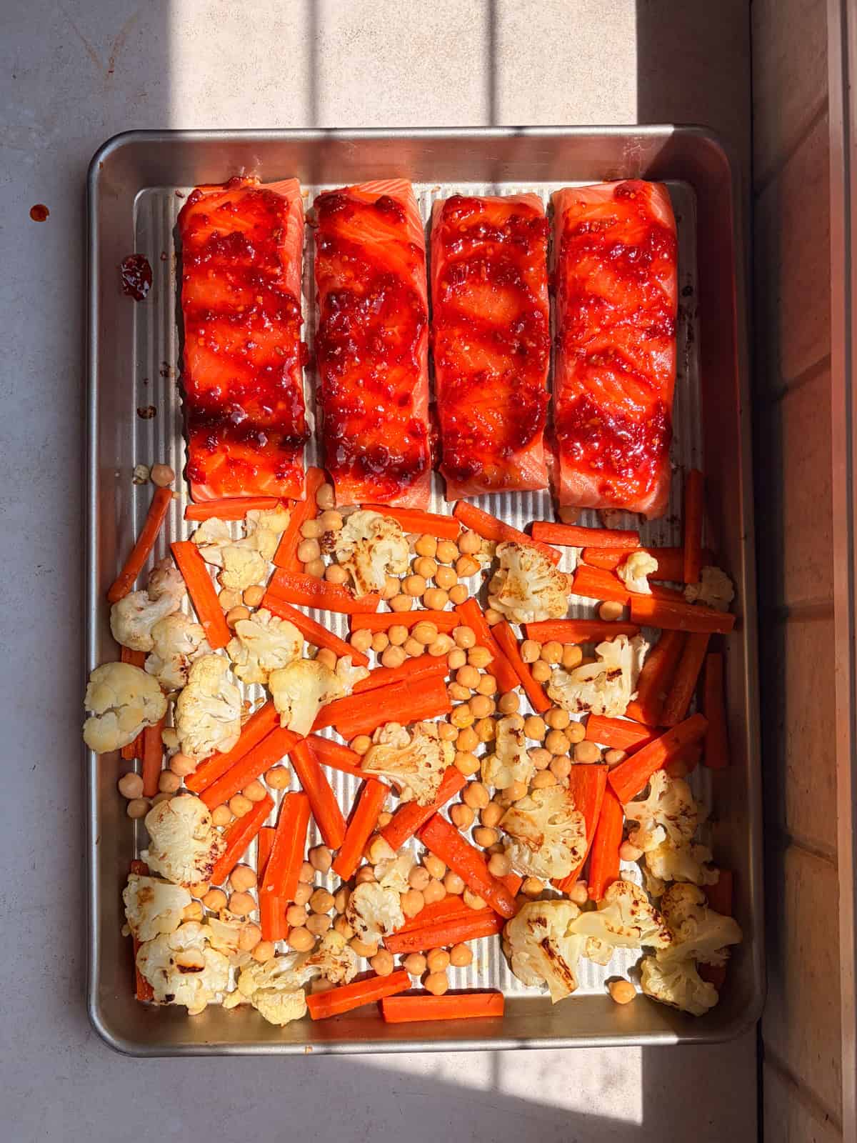 A baking tray with four harissa salmon fillets, roasted cauliflower, carrot sticks, and chickpeas—all arranged neatly and lit by sunlight.