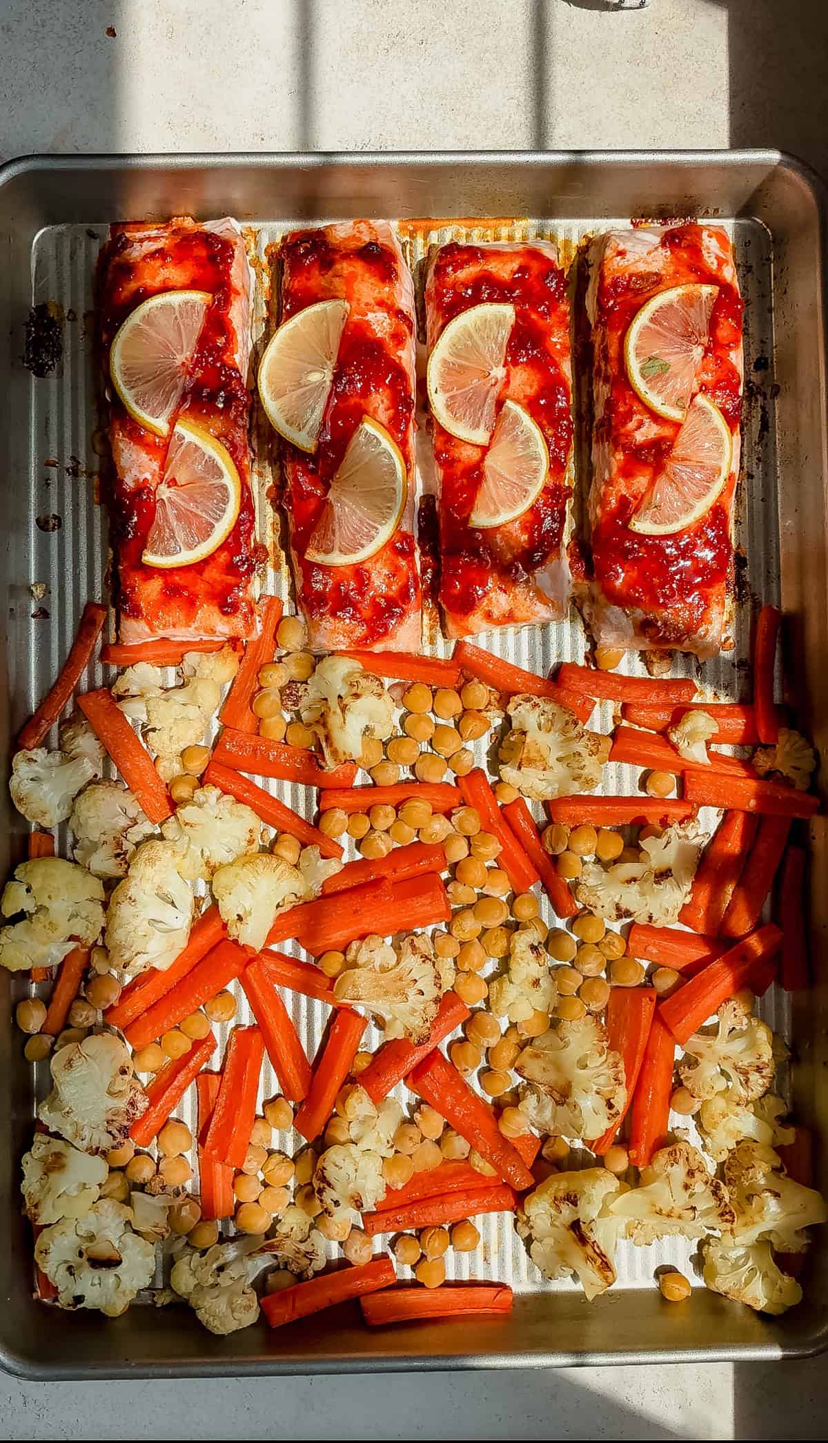 A baking sheet with four harissa salmon fillets topped with lemon slices alongside roasted carrots, cauliflower florets, and chickpeas. Sunlight casts shadows over the colorful, cooked ingredients.
