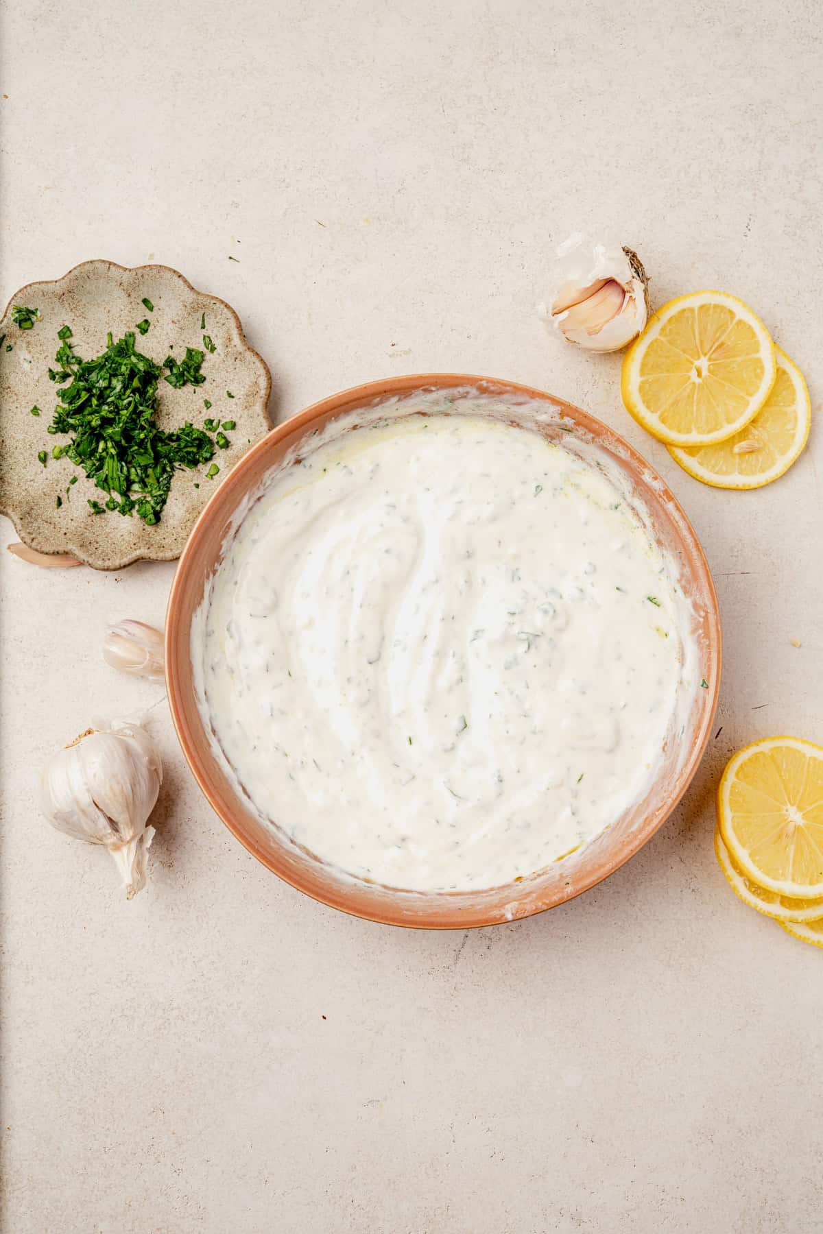 A bowl of creamy yogurt sauce with herbs sits on a light surface, surrounded by garlic, lemon slices, and a small plate of chopped parsley—the perfect accompaniment to harissa salmon.