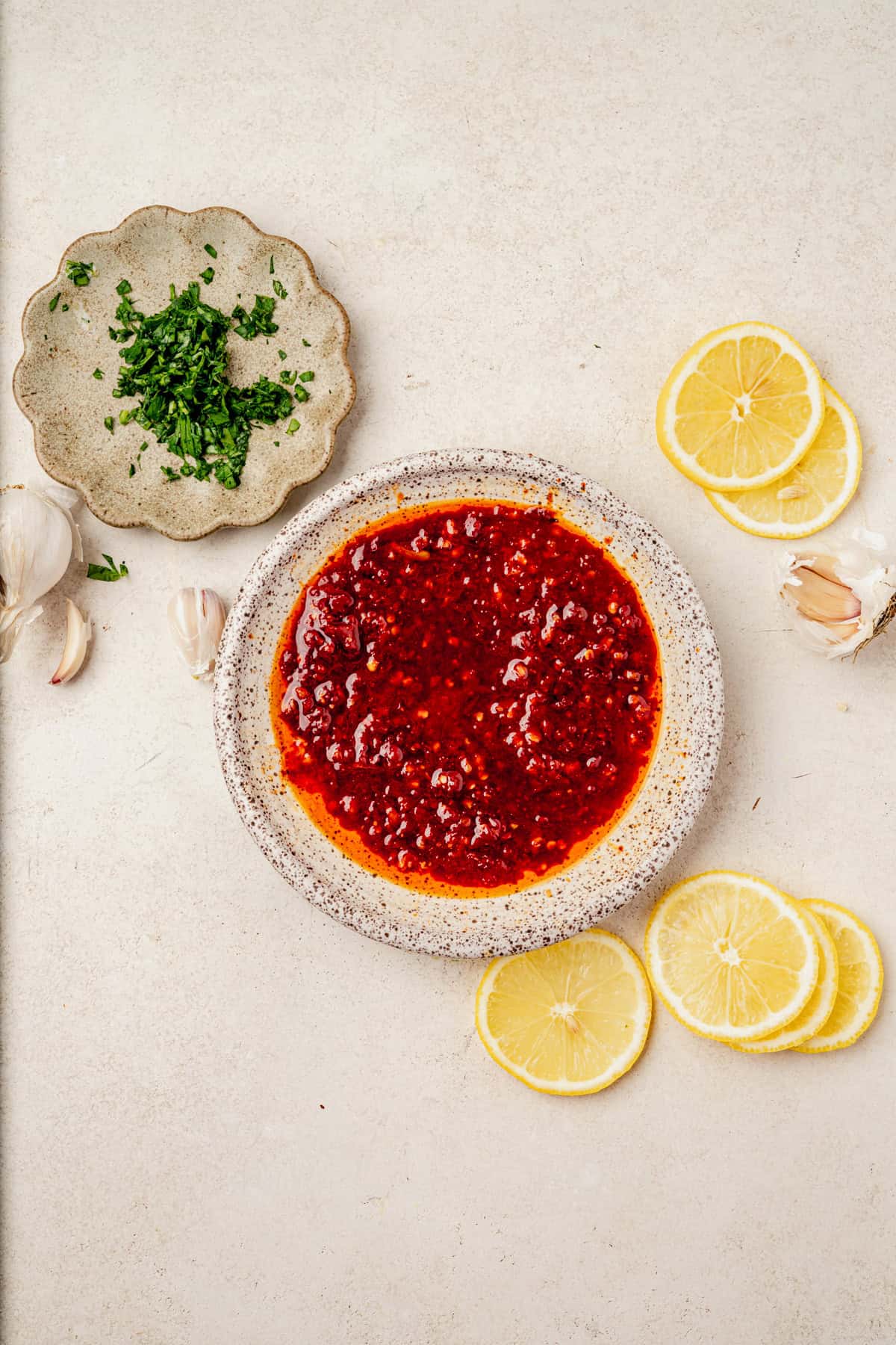 A bowl of honey harissa glaze perfect for harissa salmon, along with three lemon slices, garlic cloves, and a small plate of chopped herbs are arranged on a light surface.