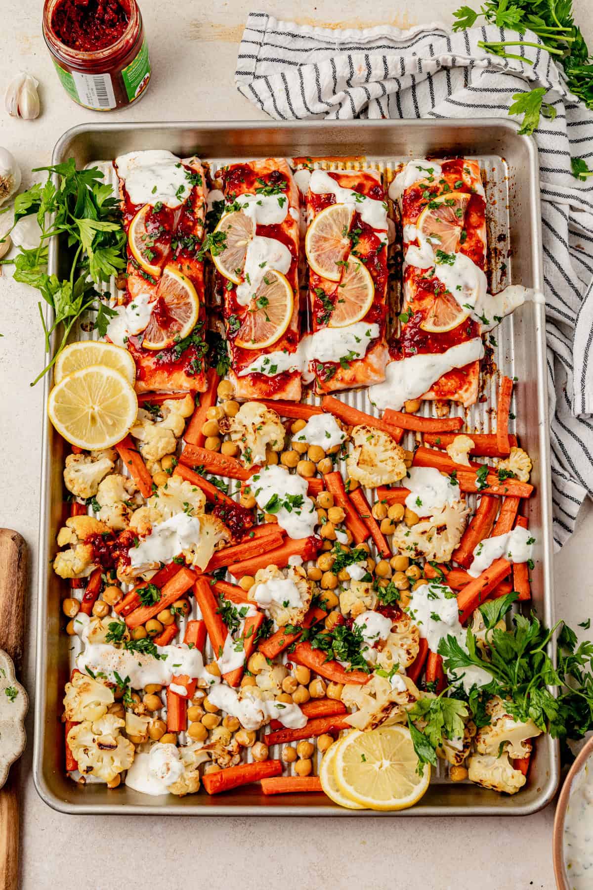 Sheet pan with harissa salmon baked to perfection, topped with lemon slices and creamy sauce, surrounded by roasted carrots, cauliflower, chickpeas, parsley, and lemon wedges. A striped towel and jar of sauce are in the background.