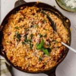 Creamy sun dried tomato orzo topped with grated cheese and fresh herbs in a cast iron skillet. A serving spoon rests in the dish, and a green plate with extra cheese is nearby.