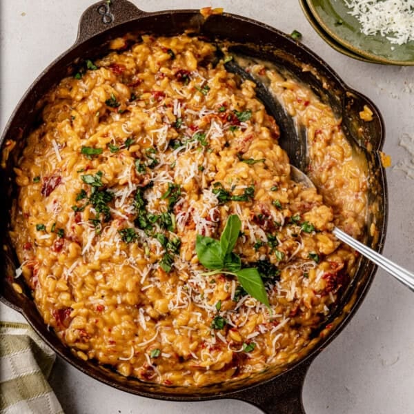Creamy sun dried tomato orzo topped with grated cheese and fresh herbs in a cast iron skillet. A serving spoon rests in the dish, and a green plate with extra cheese is nearby.