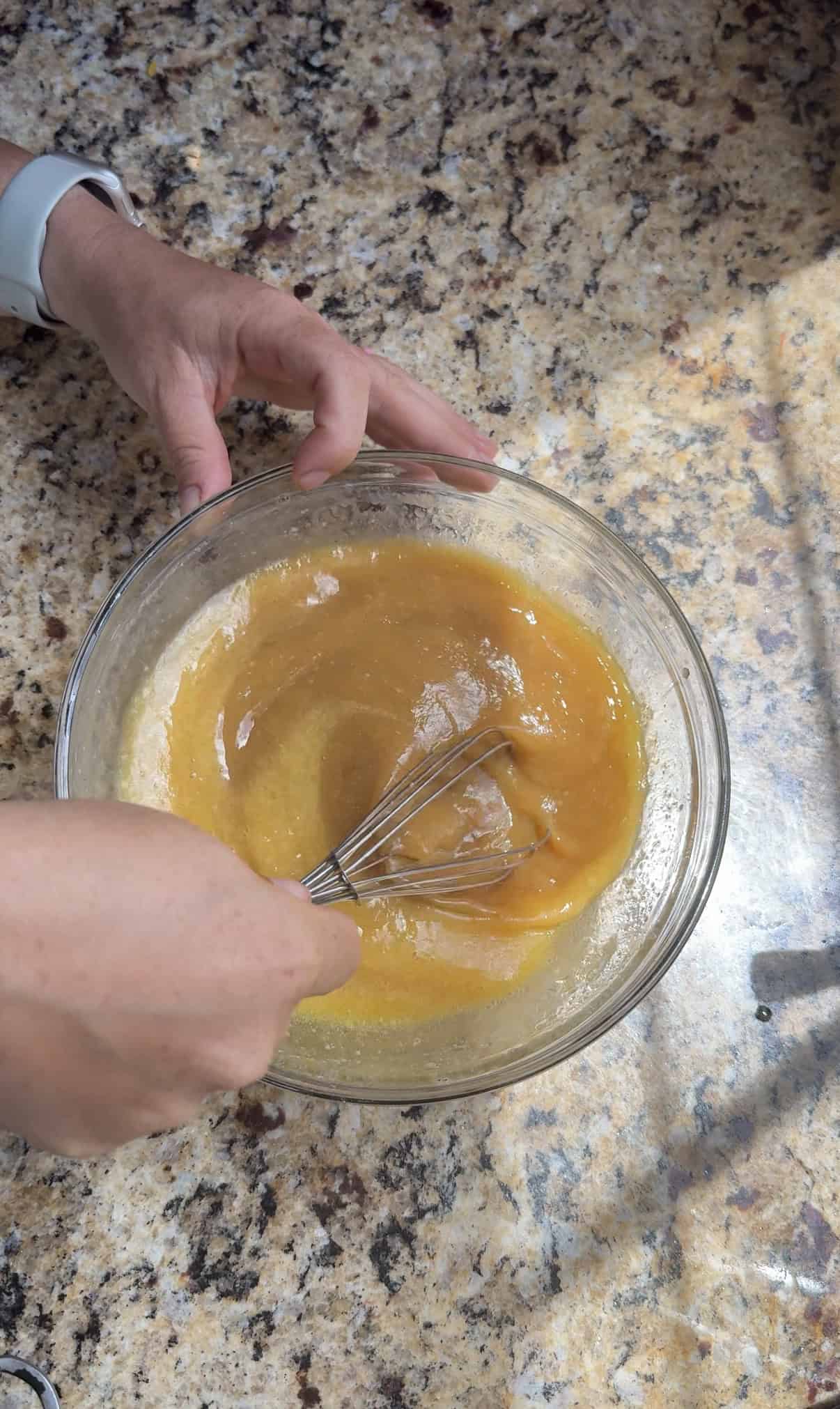 A person whisking a the wet ingredients for zucchini carrot muffins  in a clear glass bowl on a marbled countertop, with sunlight casting shadows across the surface.