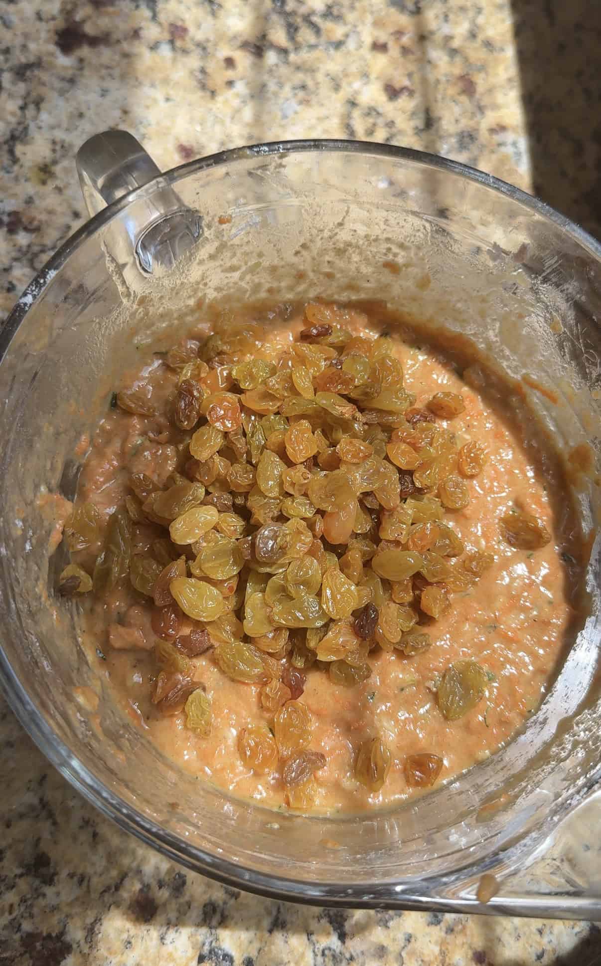 A glass bowl of zucchini carrot muffin batter with a pile of golden raisins on top, sitting on a granite countertop, lit by sunlight.