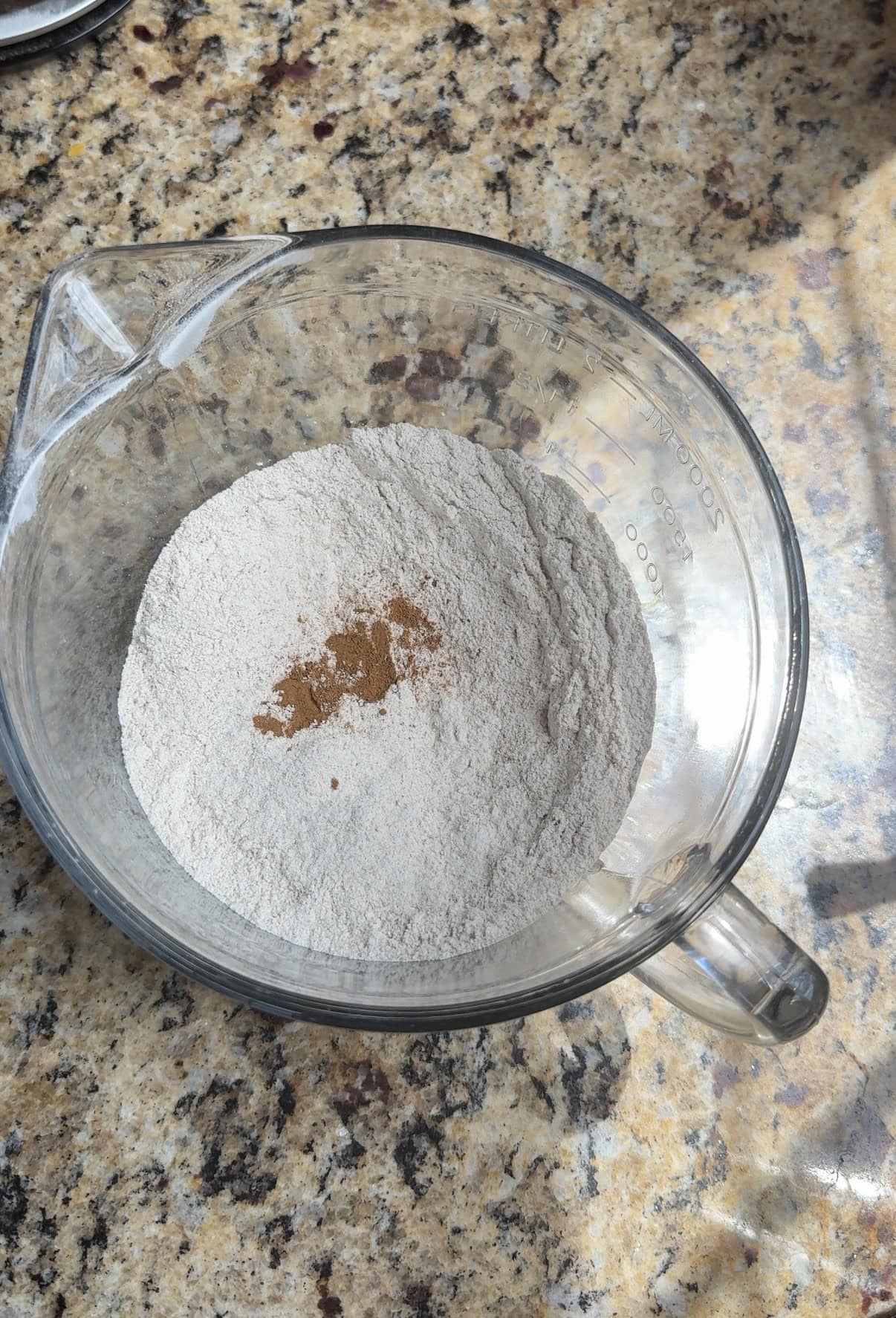 A glass mixing bowl with flour and a small amount of cinnamon on top sits on a speckled countertop, lit by sunlight.