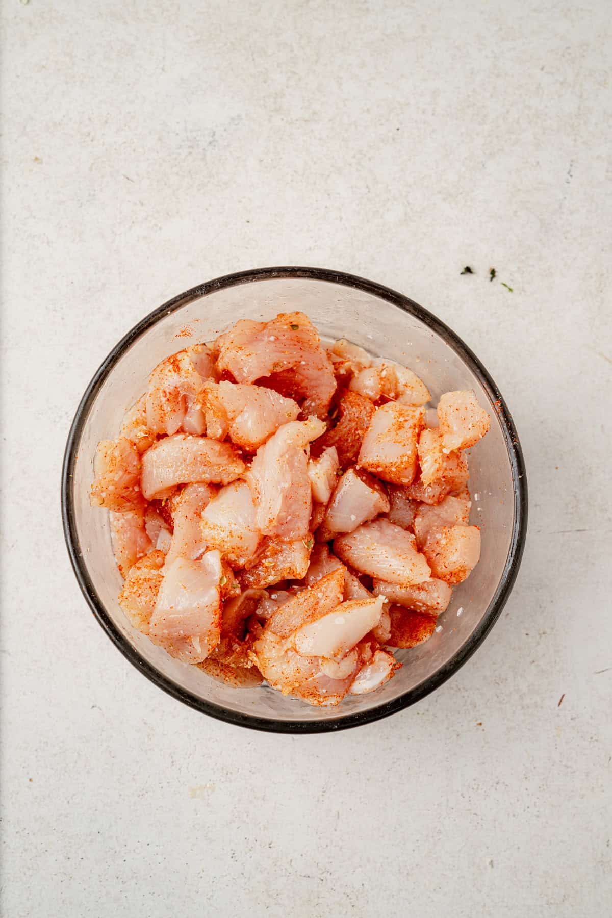 A glass bowl filled with raw, diced chicken pieces coated in seasoning, ready to be added to a flavorful chicken zucchini skillet, placed on a light-colored surface.