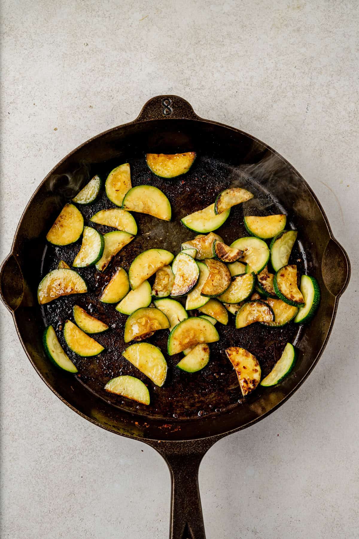 Sliced zucchini is being sautéed in a cast iron skillet with browned bits and oil, placed on a light-colored surface.