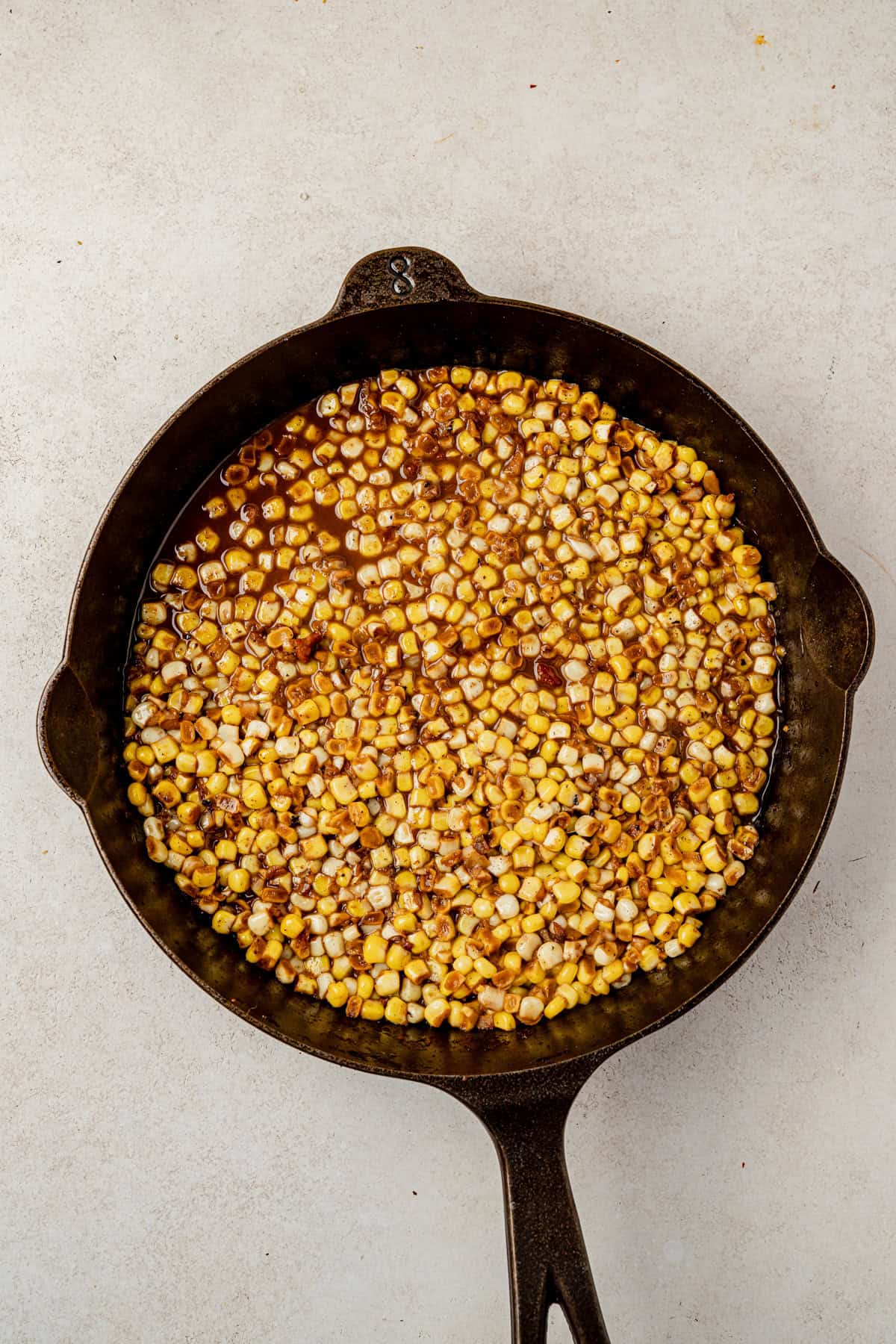 A cast iron skillet filled with sautéed corn kernels, lightly browned and glistening with a white wine pan sauce, sits on a light-colored surface.