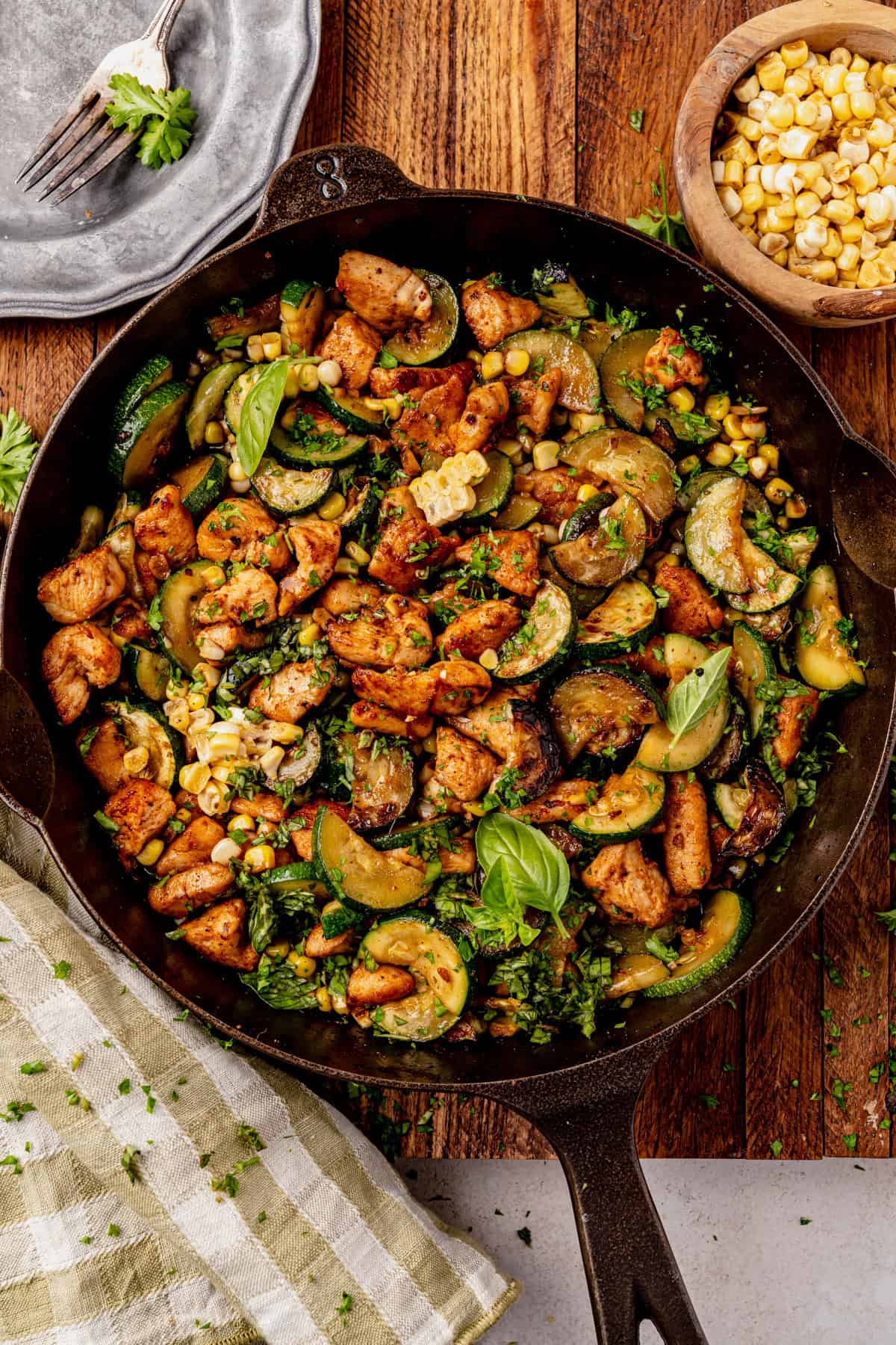 A cast iron chicken zucchini skillet filled with sautéed chicken, zucchini, corn, and herbs sits on a wooden table, with a plate, fork, napkin, and bowl of corn nearby.