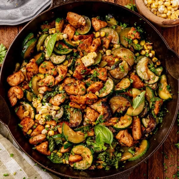 A chicken zucchini skillet filled with sautéed chicken, zucchini, corn, and fresh herbs, garnished with basil leaves, sits on a wooden table next to a small bowl of corn and a striped cloth napkin.