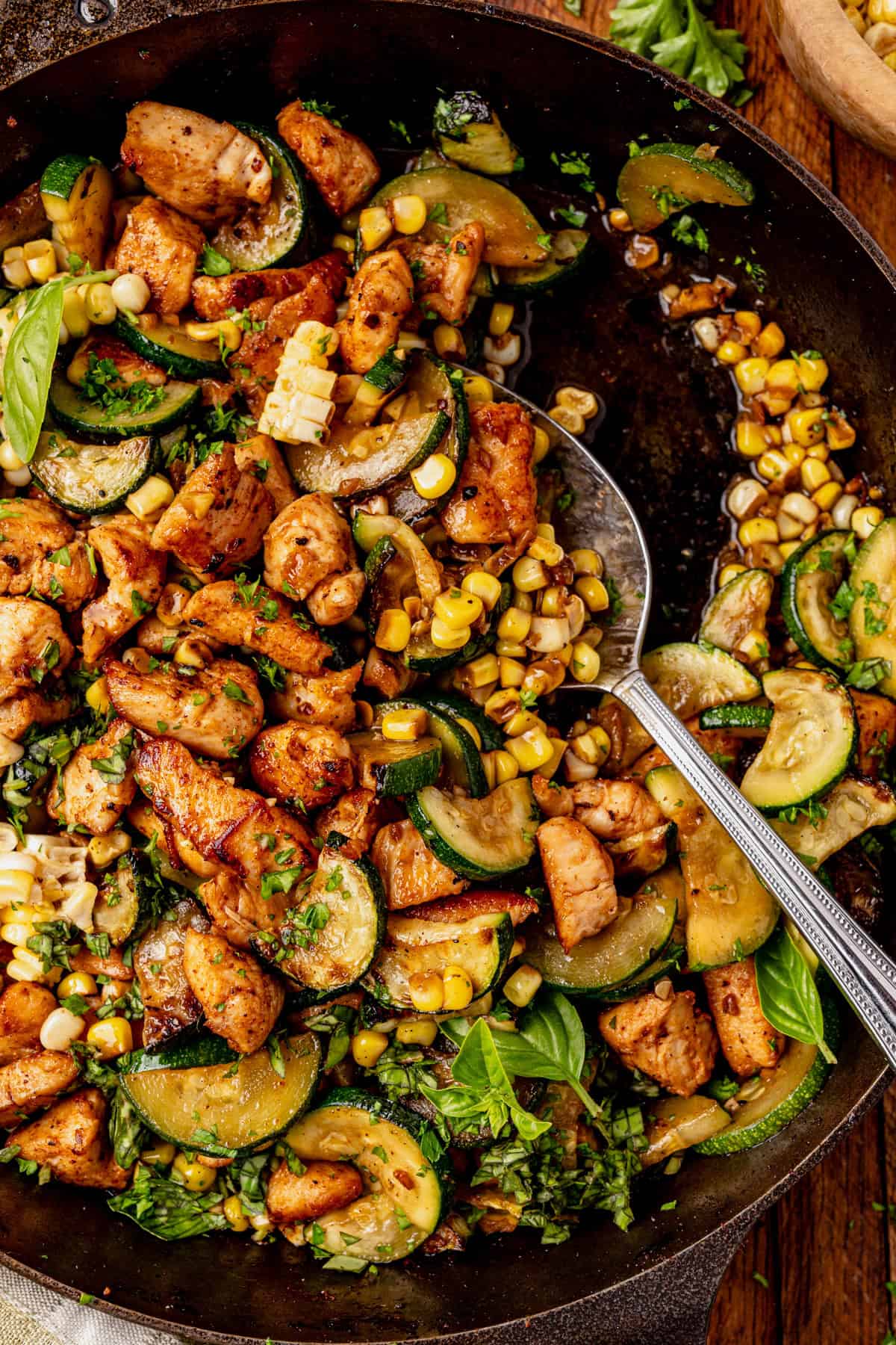 A chicken zucchini skillet filled with cooked chicken, sliced zucchini, corn, and fresh herbs sits on a wooden surface, with a spoon resting inside.