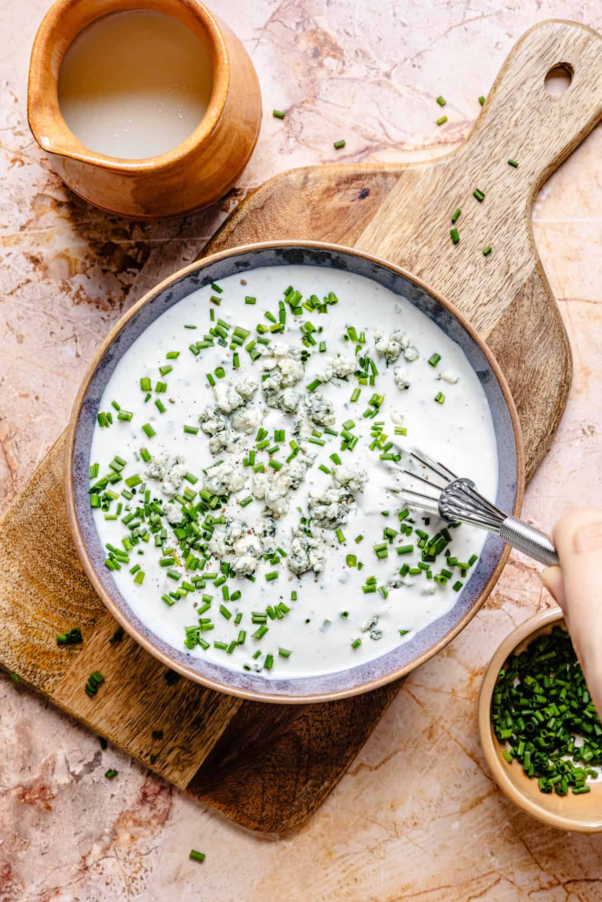 A bowl of creamy dressing with crumbled blue cheese and chopped chives sits on a wooden board, ready to be drizzled over a blue cheese and steak salad. Nearby, a small bowl of chives and a clay cup of liquid complete the rustic scene as a hand mixes the dressing with a fork.