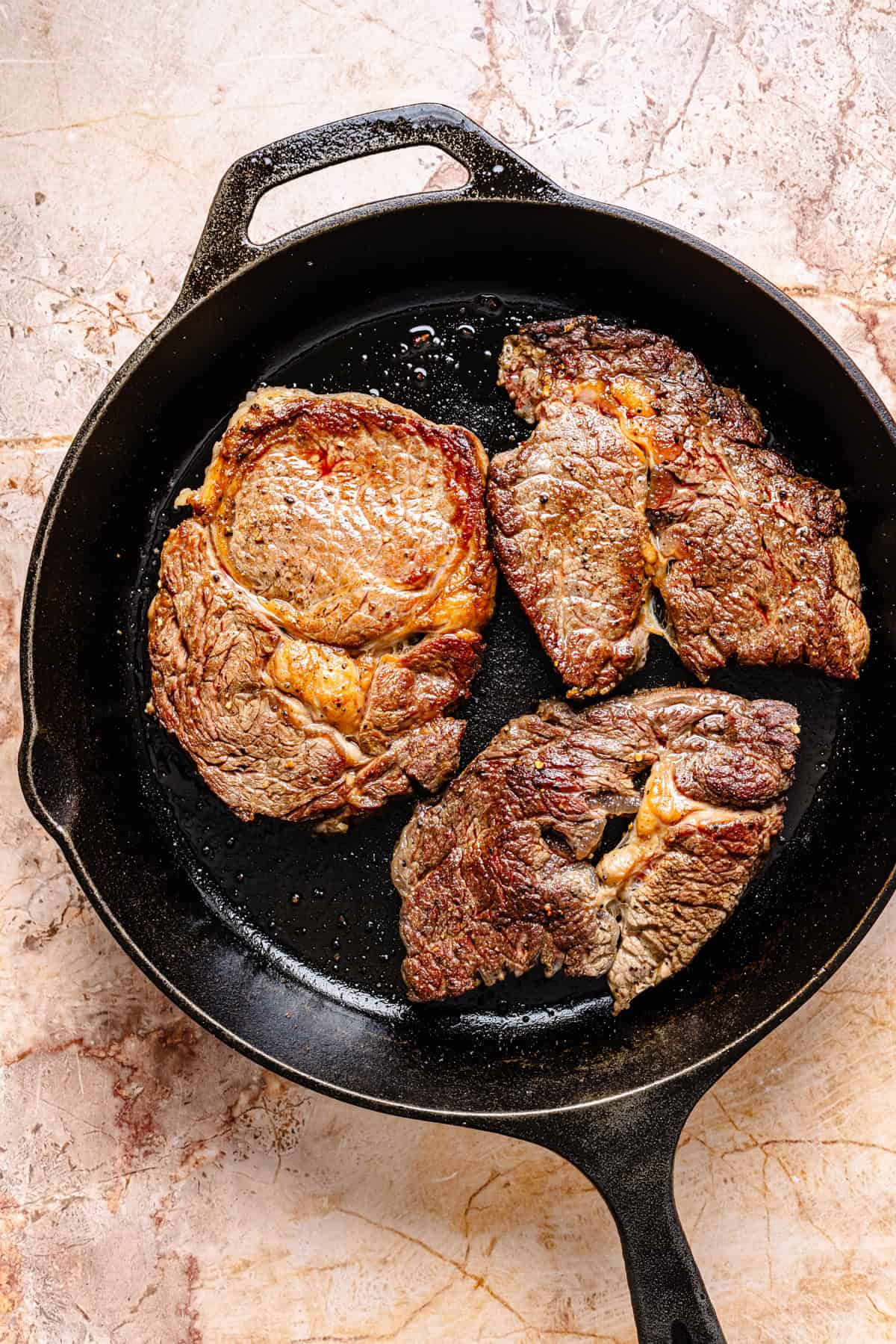Three seared steaks cooking in a black cast iron skillet on a light-colored stone countertop, perfect for slicing and serving atop a fresh blue cheese and steak salad.