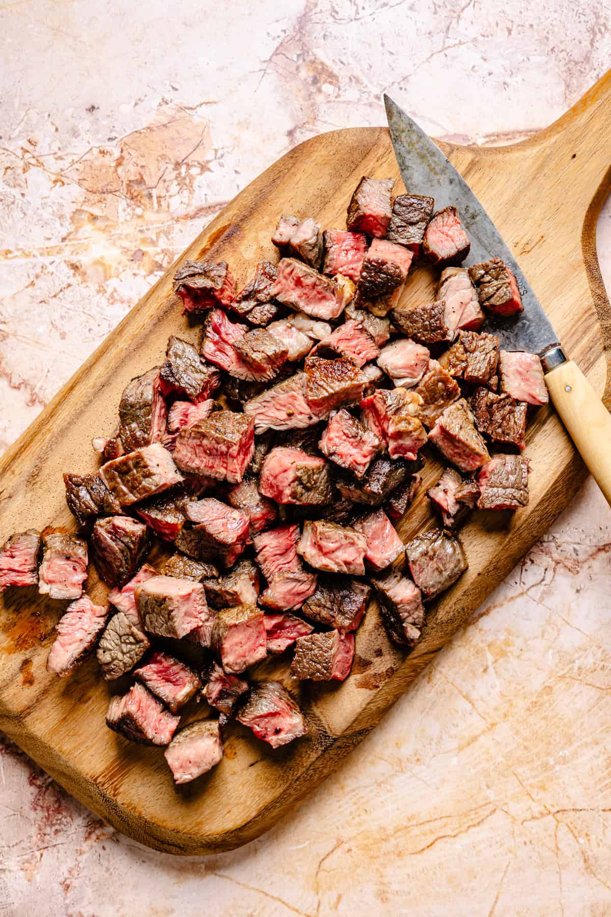 Cubed pieces of cooked steak with a seared exterior and pink center are scattered on a wooden cutting board next to a knife with a light-colored handle, ready to be tossed into a fresh blue cheese and steak salad.