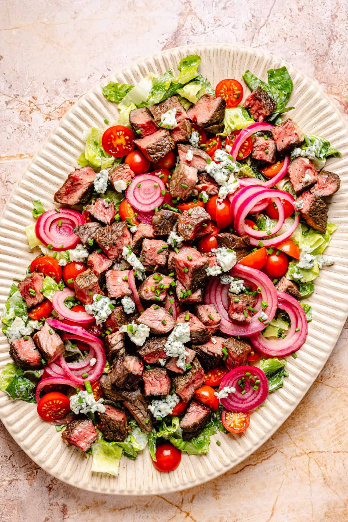 Oval platter with chopped steak, blue cheese crumbles, pickled red onions, cherry tomatoes, and Romaine lettuce—everything you need for a flavorful blue cheese and steak salad—artfully arranged on a light marble surface.