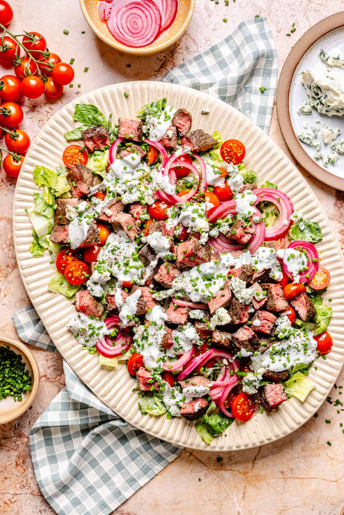 A large platter of blue cheese and steak salad with chopped lettuce, cherry tomatoes, pickled onions, blue cheese crumbles, chives, and creamy dressing is surrounded by bowls of ingredients and a blue-checkered napkin.