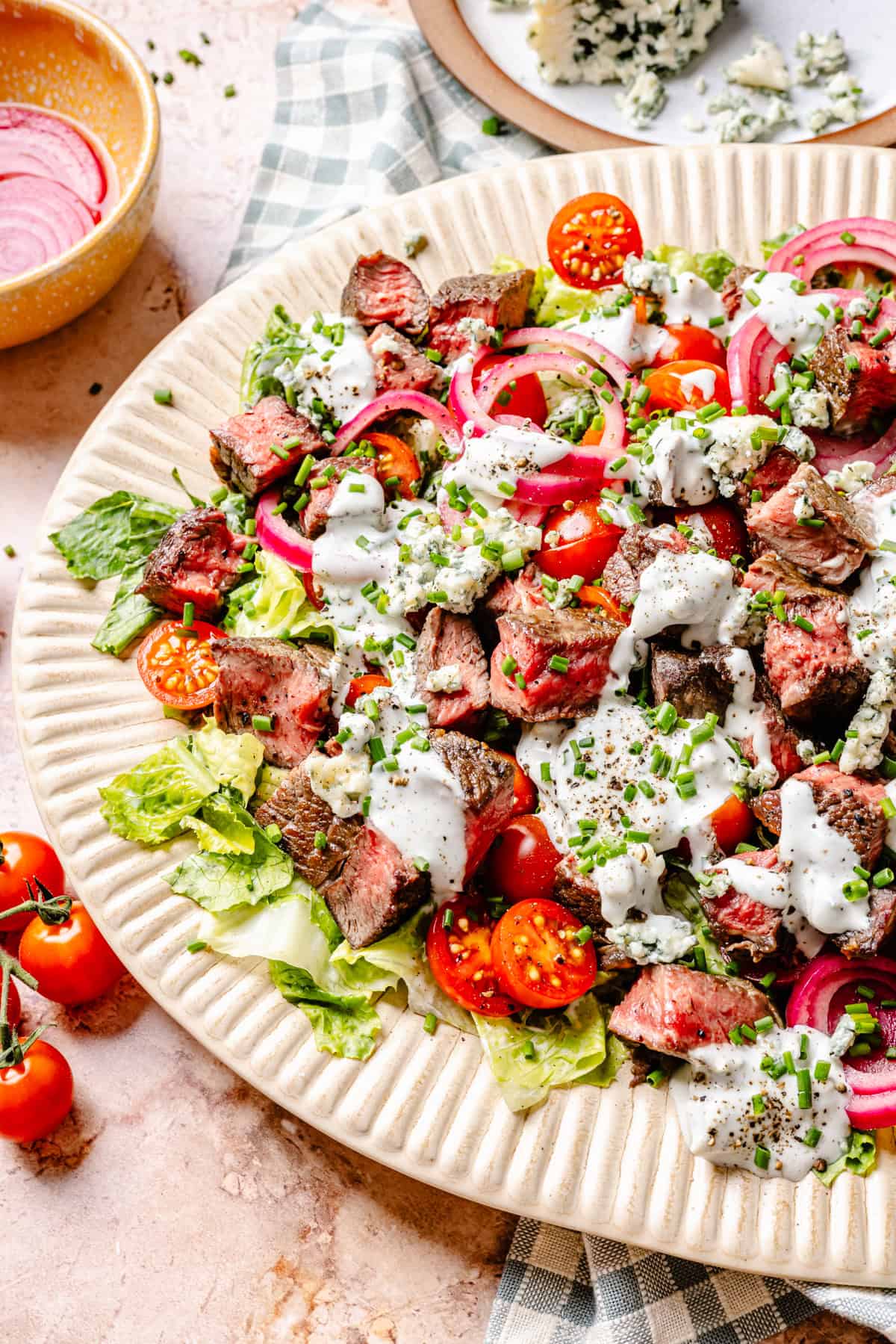 A plate of blue cheese and steak salad with chopped lettuce, cherry tomatoes, pickled onions, blue cheese crumbles, and creamy dressing, garnished with chives. A bowl of onions and a cheese plate are in the background.