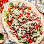 A platter of blue cheese and steak salad with chopped lettuce, cherry tomatoes, red onions, blue cheese crumbles, chives, and creamy dressing. Blue cheese, chives, and tomatoes are served on the side.