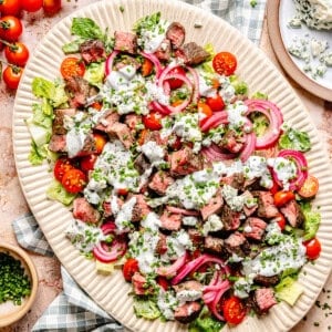 A platter of blue cheese and steak salad with chopped lettuce, cherry tomatoes, red onions, blue cheese crumbles, chives, and creamy dressing. Blue cheese, chives, and tomatoes are served on the side.