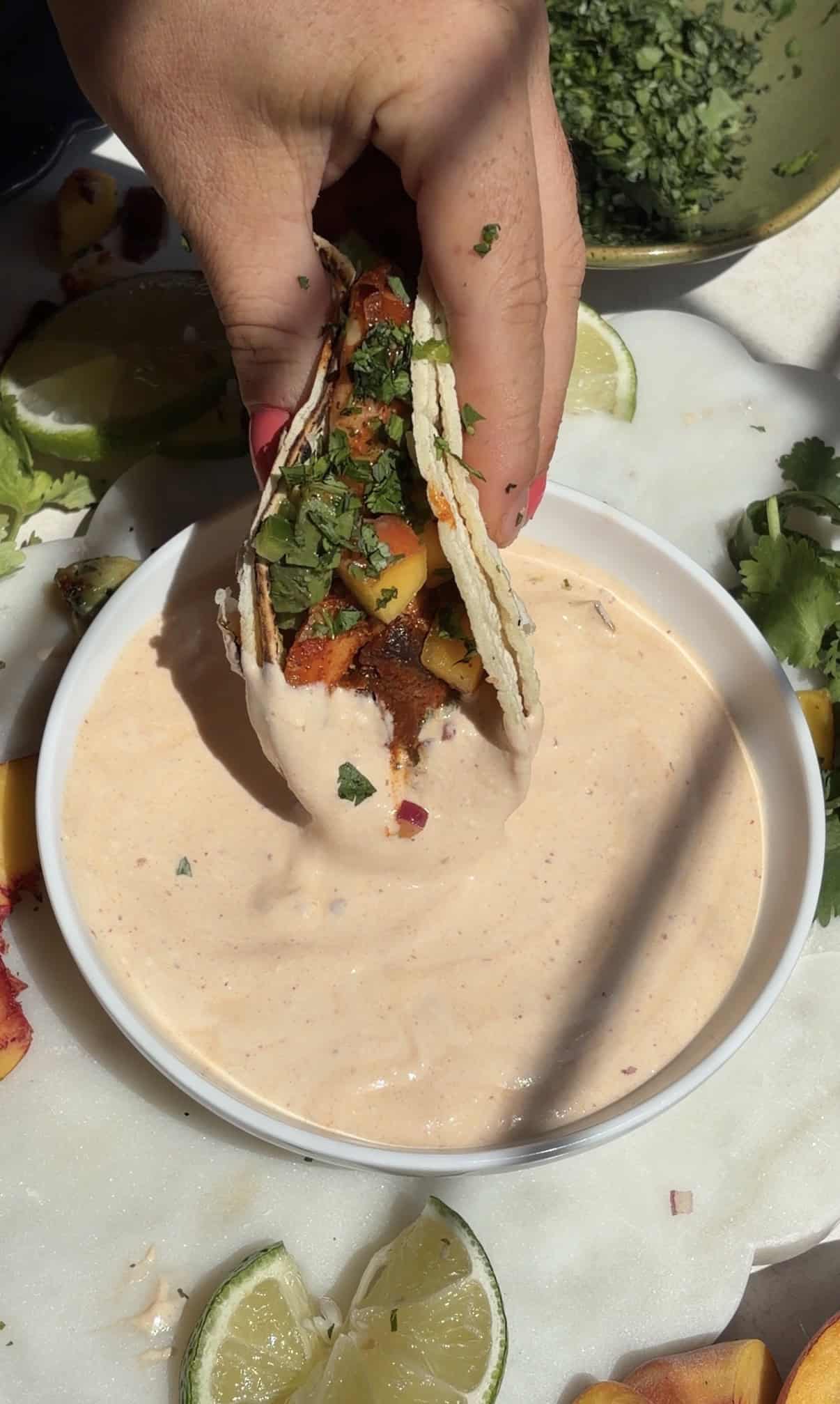 A hand holds a chipotle shrimp taco filled with vegetables and herbs, dipping it into a bowl of creamy sauce. Lime wedges and chopped cilantro are visible on the white surface nearby.