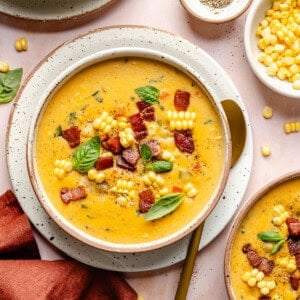 Creamy corn chowder garnished with bacon, fresh basil, and corn kernels in a white bowl, with a gold spoon beside it. Bowls of black pepper and extra corn are nearby on a peach-colored surface.