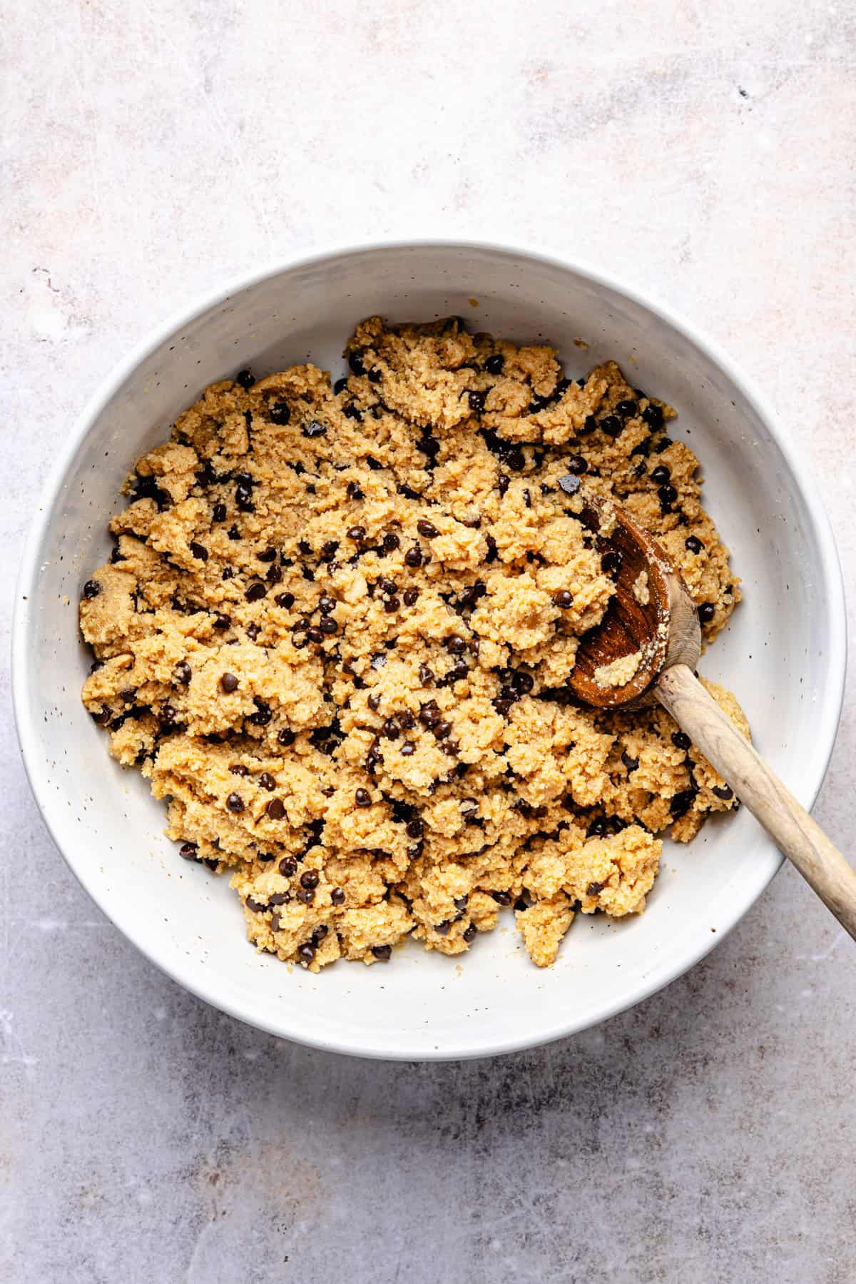 A white bowl filled with chocolate chip protein cookie dough, mixed with a wooden spoon, sits on a light-colored countertop—perfect for making delicious protein cookie dough bark.