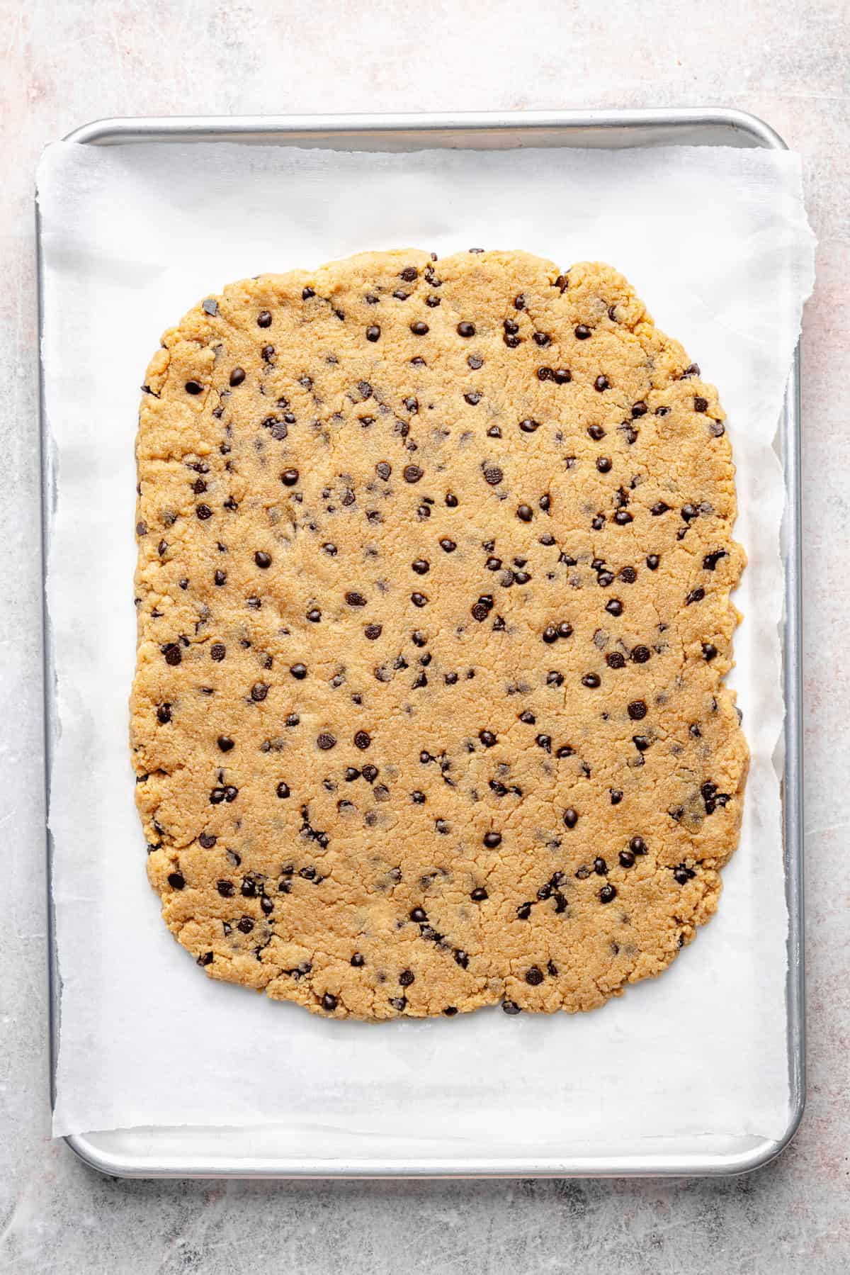A slab of protein cookie dough bark is spread out on a parchment-lined baking sheet, ready to be baked.