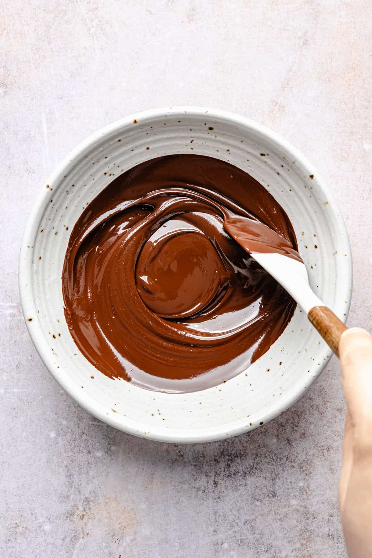 A hand uses a spatula to stir glossy melted chocolate in a white speckled ceramic bowl on a light countertop, preparing the base for delicious protein cookie dough bark.