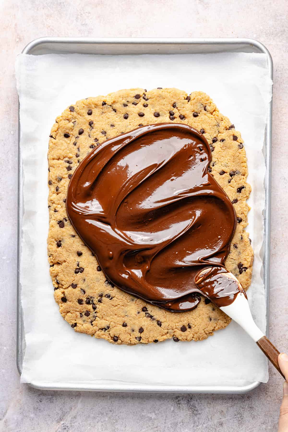 A large rectangular protein cookie dough bark with chocolate chips on a baking tray is being spread with melted chocolate using a spatula.