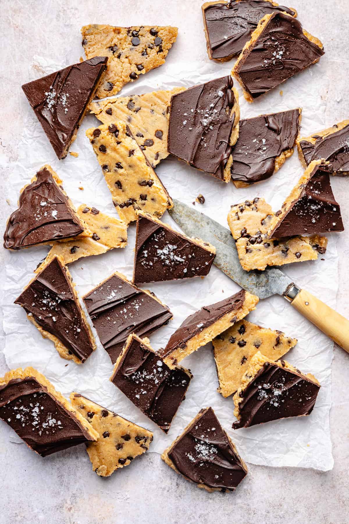 Chocolate-covered protein cookie dough bark bars are scattered on parchment paper, some sprinkled with sea salt. A knife with a wooden handle lies among the bars. The treats are triangular and irregularly shaped.