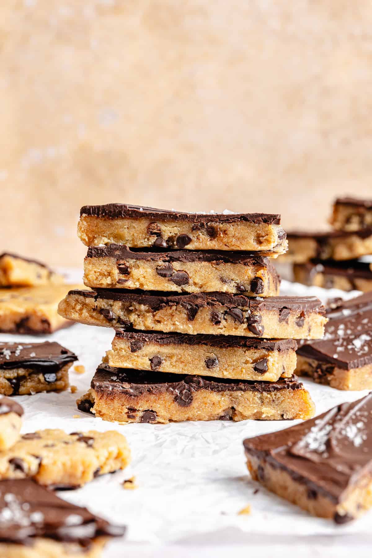 A stack of chocolate chip cookie protein bark topped with a layer of chocolate, surrounded by more bars and crumbs on white parchment paper.