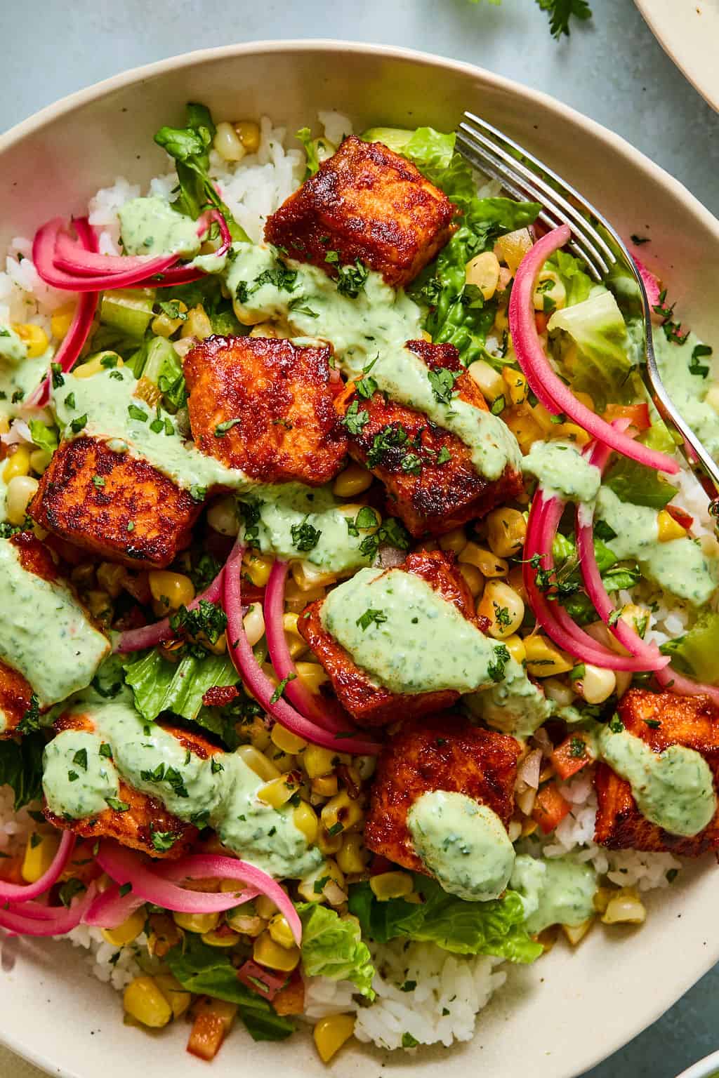 A bowl with rice, grilled salmon cubes, lettuce, corn, pickled red onions, chopped herbs, and creamy green sauce—this dish captures all the fresh flavors of salmon taco bowls—served with a fork on the side.