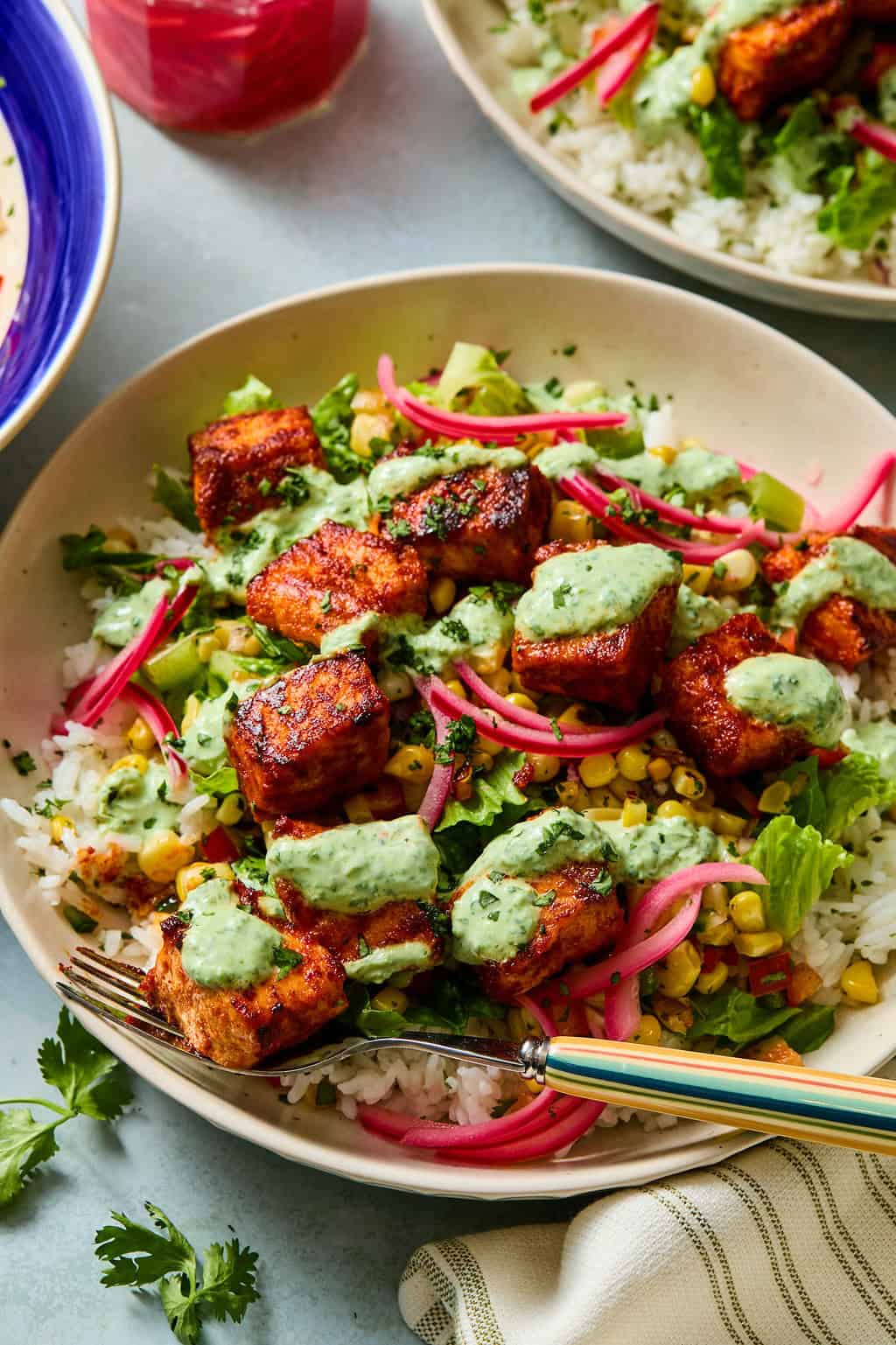 A plate of rice topped with crispy taco salmon cubes, corn salsa, lettuce, pickled onions, and green sauce, served with a fork and garnished with fresh herbs.