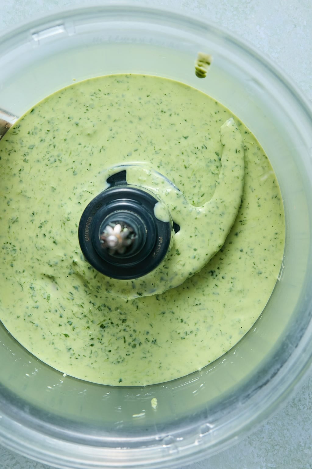A food processor bowl filled with a creamy, green herb sauce, with flecks of herbs visible throughout—perfect for drizzling over salmon taco bowls. The food processor blade is in the center, and the sauce has a smooth, thick texture.