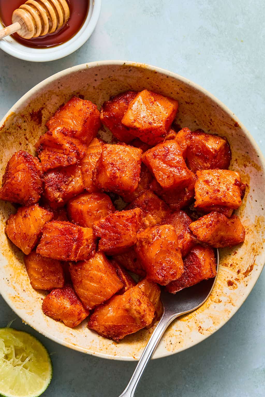 A bowl of seasoned, cooked salmon bites with a spoon, next to a lime wedge and a small dish of honey with a dipper—perfect for building your own salmon taco bowls.
