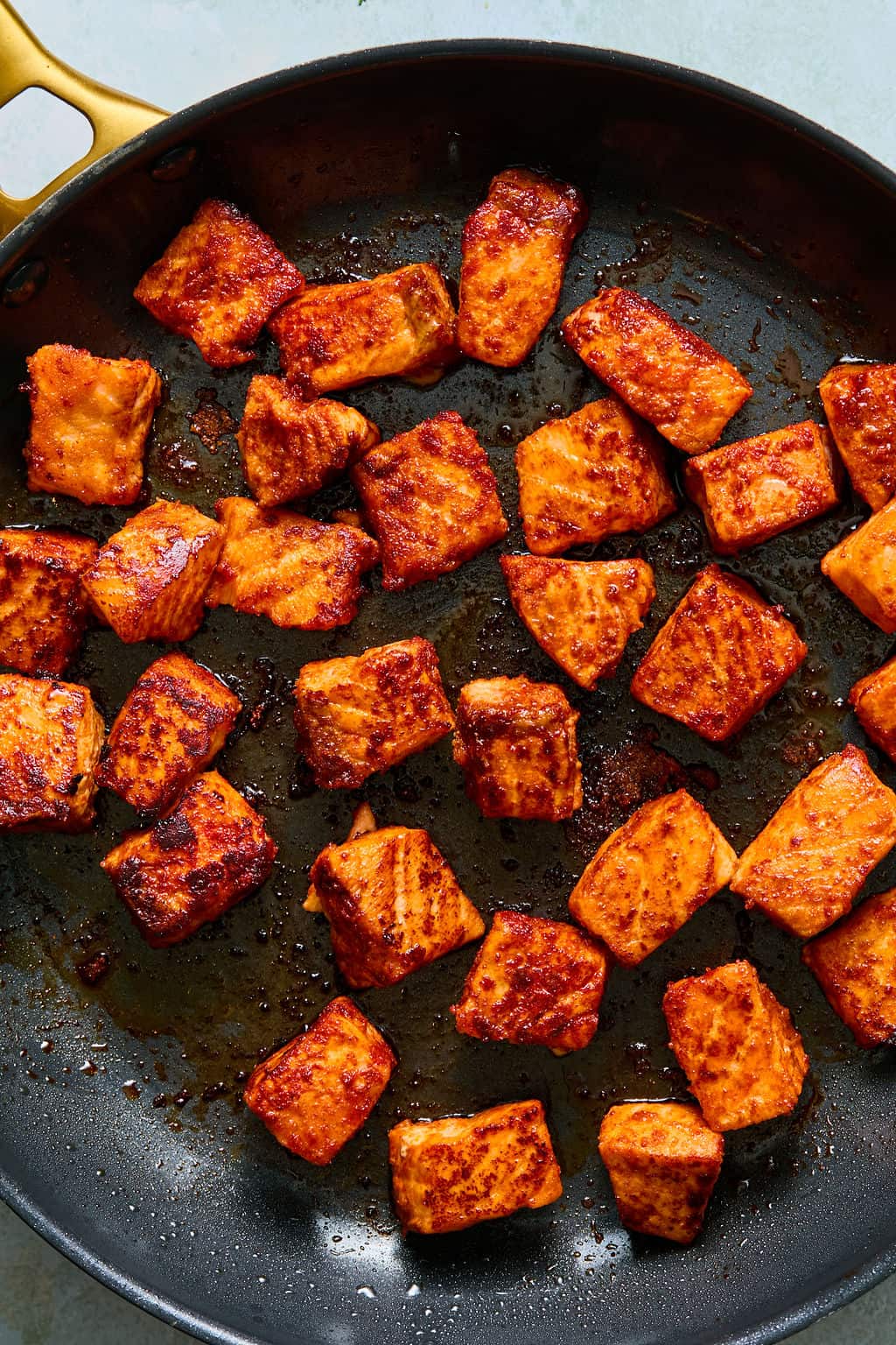 Cubed pieces of salmon coated with spices are being pan-seared in a nonstick skillet, creating a crispy, golden-brown exterior—perfect for assembling flavorful salmon taco bowls. The pan has a gold handle and is shown from above.