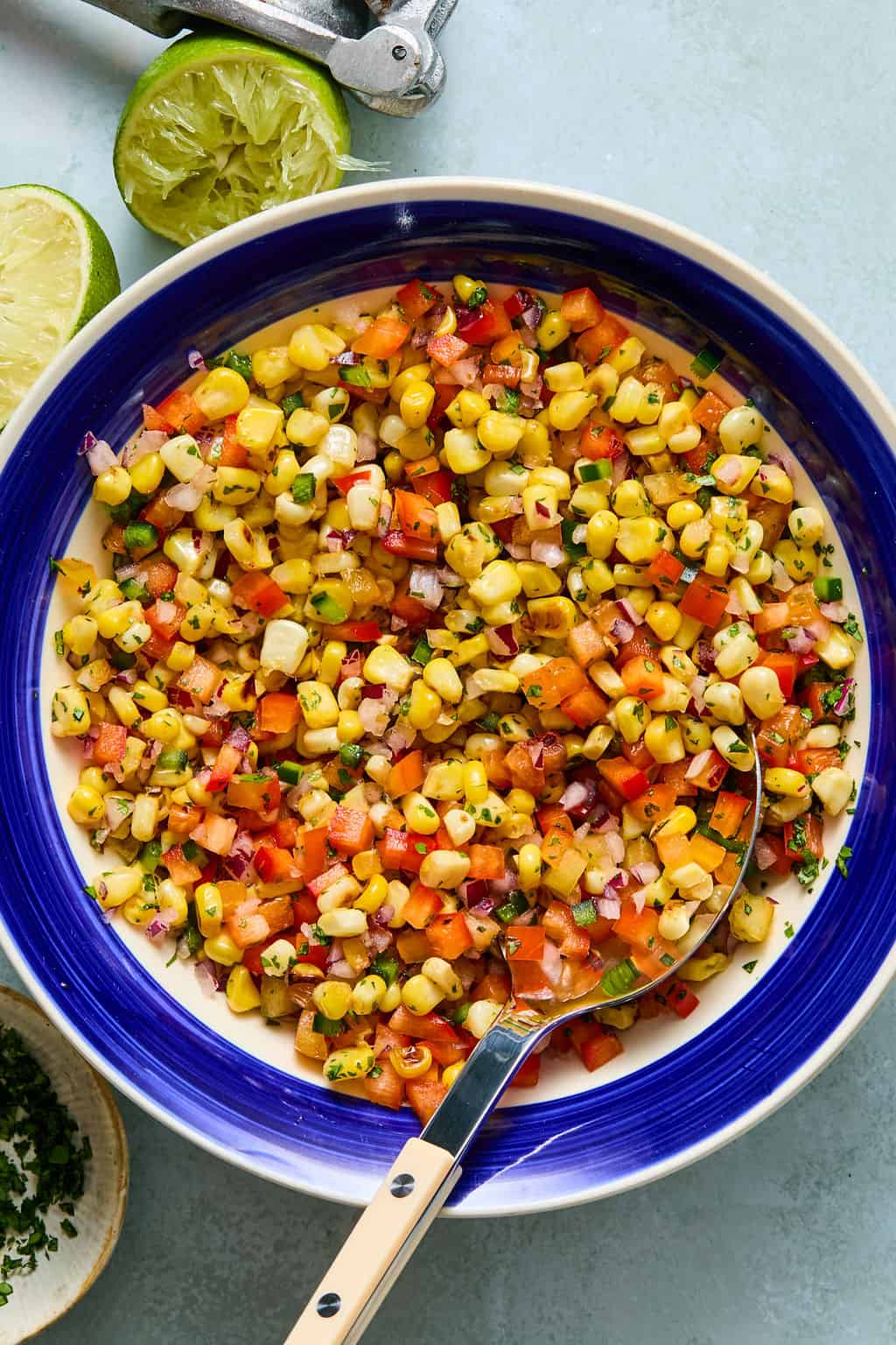 A bowl of corn salsa with diced tomatoes, onions, herbs, and peppers, with a spoon in it—perfect as a fresh topping for salmon taco bowls. Lime halves and a citrus squeezer are nearby on the table.