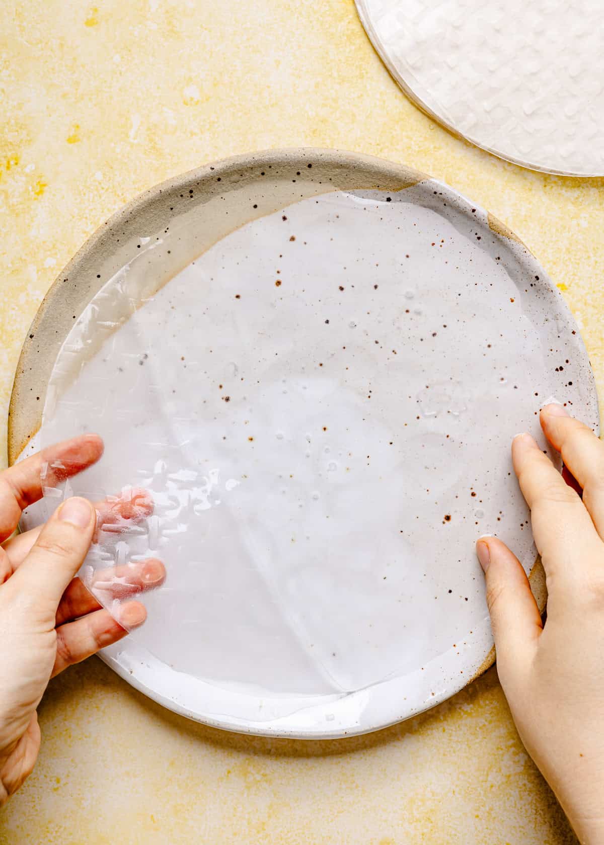 Two hands hold a wet rice paper sheet on a speckled ceramic plate, preparing the base for Thai summer rolls with peanut sauce, with another dry rice paper sheet visible nearby, all on a yellow textured surface.
