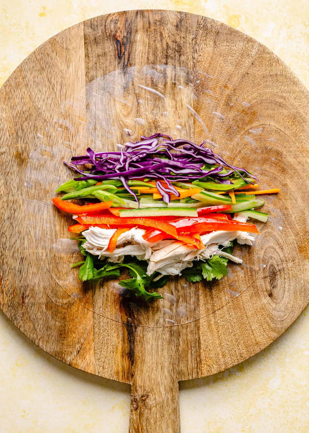 Assorted fresh vegetables and shredded chicken are neatly arranged in rows on a round wooden board, ready to be wrapped for vibrant Thai summer rolls with peanut sauce, with cilantro, red pepper, chicken, carrots, green pepper, and purple cabbage visible.