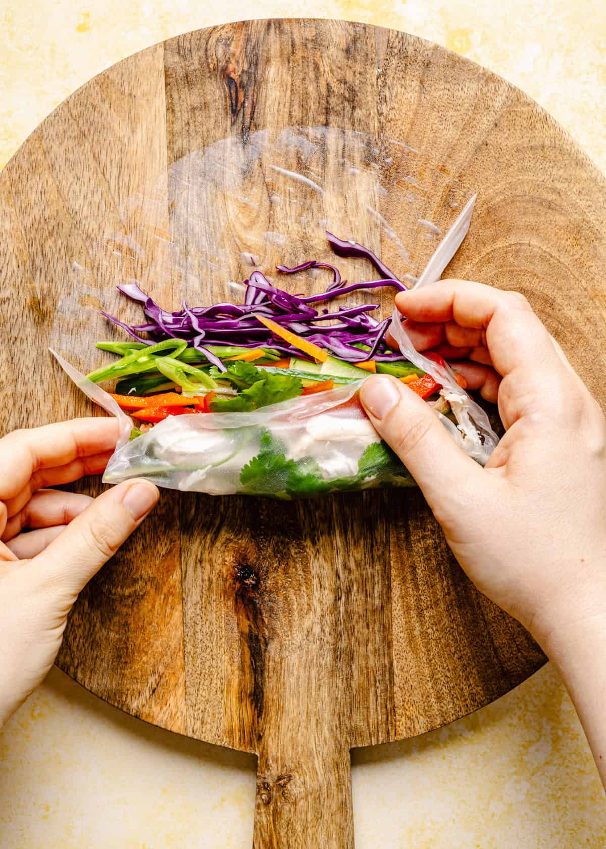 Two hands are rolling a fresh Thai summer roll with rice paper, red cabbage, carrots, cilantro, and other vegetables on a round wooden cutting board—perfect for pairing with creamy peanut sauce.