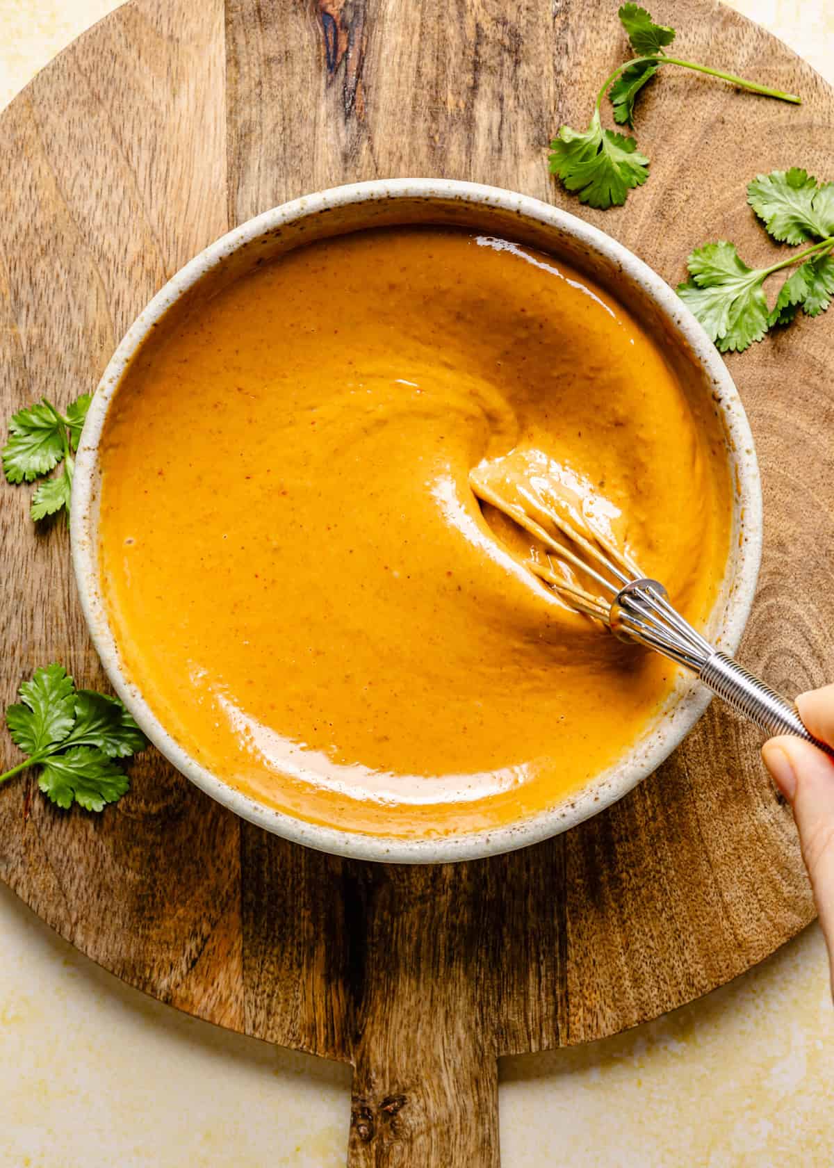A hand whisks a bowl of creamy orange sauce—perfect for thai summer rolls with peanut sauce—on a wooden board, surrounded by fresh cilantro leaves.
