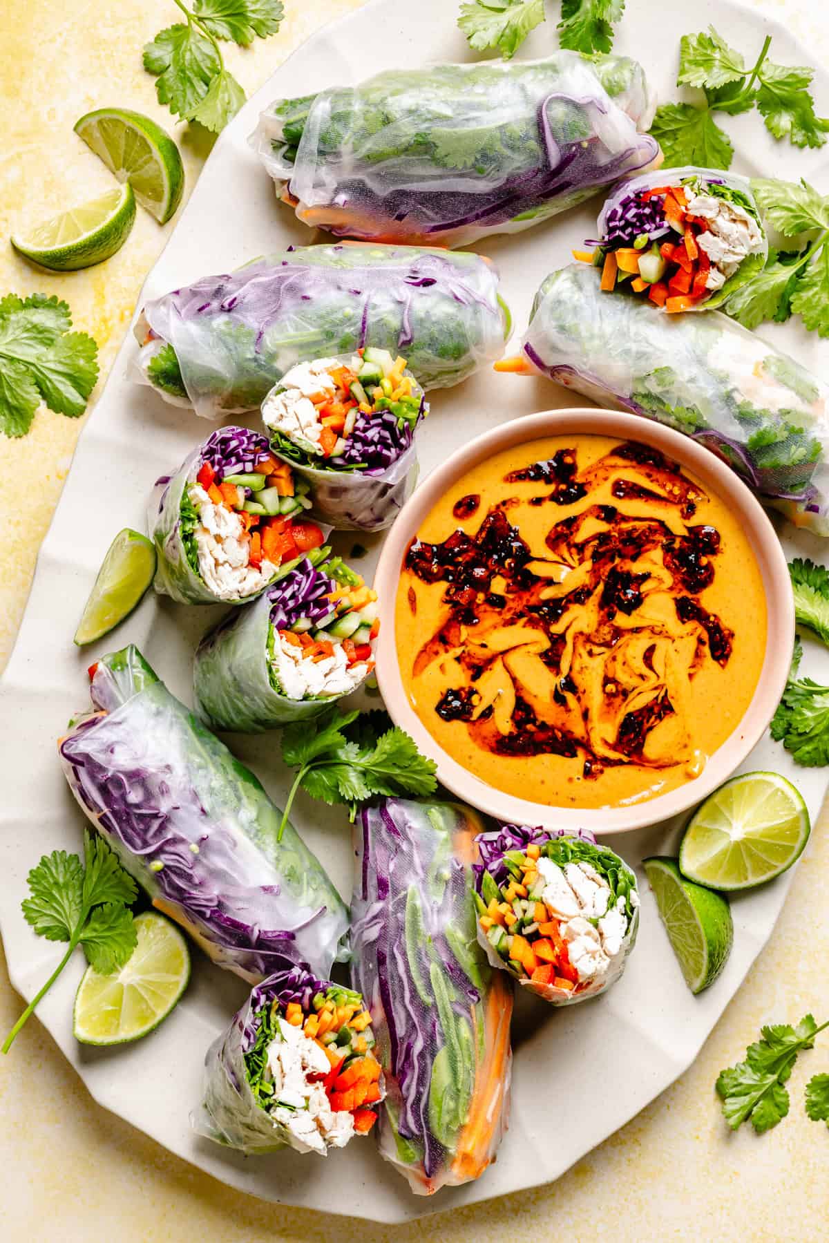 Fresh vegetable spring rolls with purple cabbage, carrots, bell peppers, and herbs, arranged around a bowl of orange chili oil dipping sauce. These Thai summer rolls with peanut sauce are garnished with lime wedges and cilantro on a white platter.