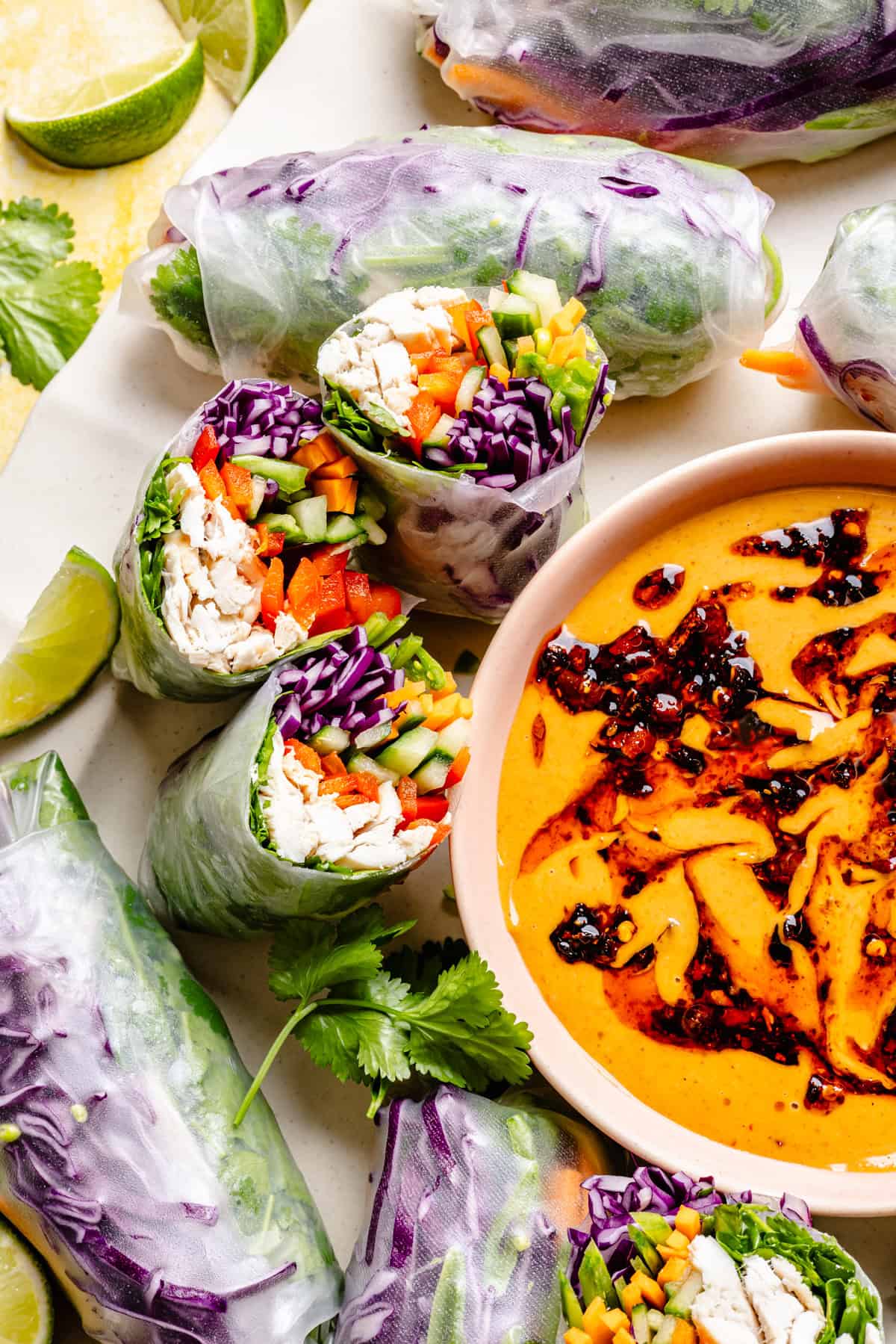 Fresh thai summer rolls with peanut sauce filled with chicken, red cabbage, cucumber, and red pepper are arranged next to a bowl of creamy orange dipping sauce with chili oil. Lime wedges and cilantro are scattered around.