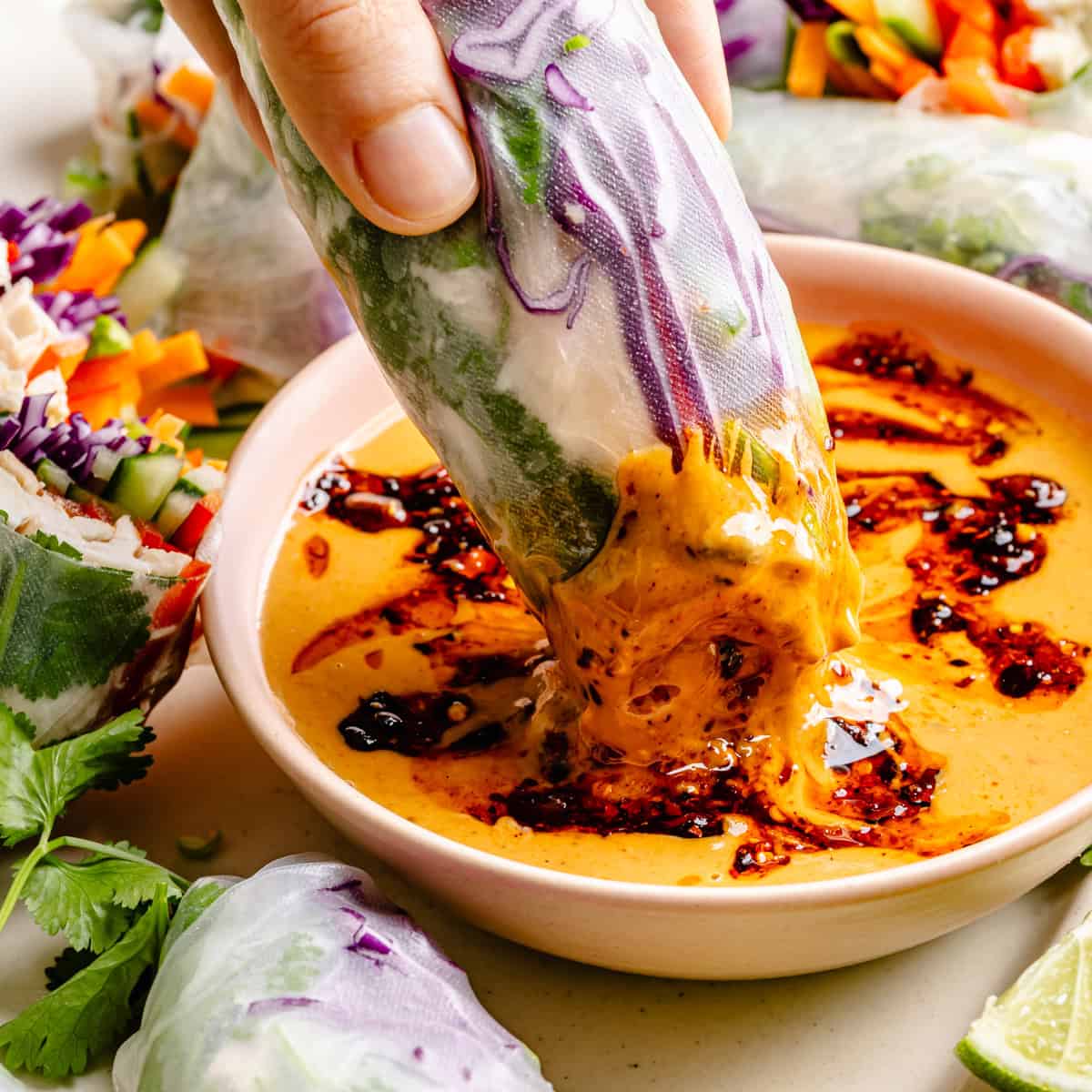 A hand dips a fresh thai summer roll with peanut sauce—packed with crisp veggies—into a creamy, spicy orange dipping sauce in a small bowl. Other spring rolls and leafy greens sit nearby on the table.