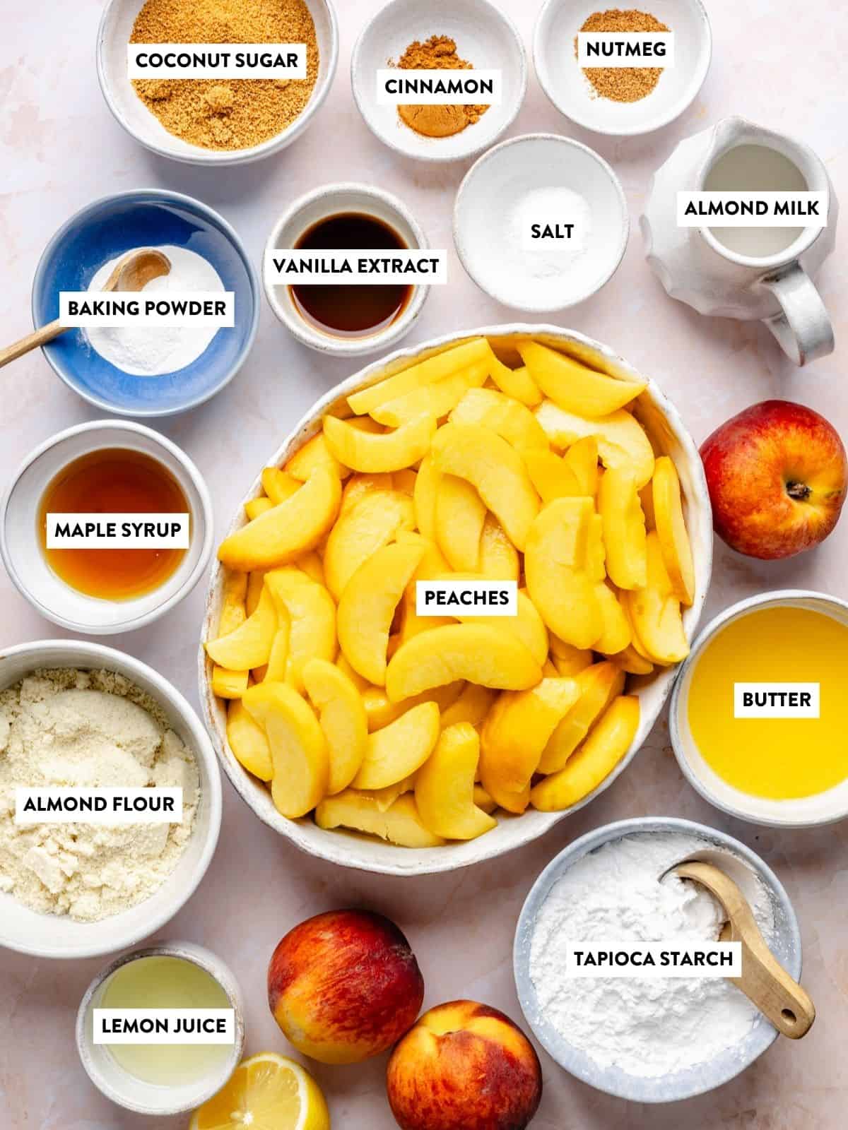A flat lay of labeled ingredients for a peach dish, including peaches, almond flour, tapioca starch, almond milk, butter, vanilla, lemon juice, maple syrup, baking powder, coconut sugar, cinnamon, nutmeg, and salt.