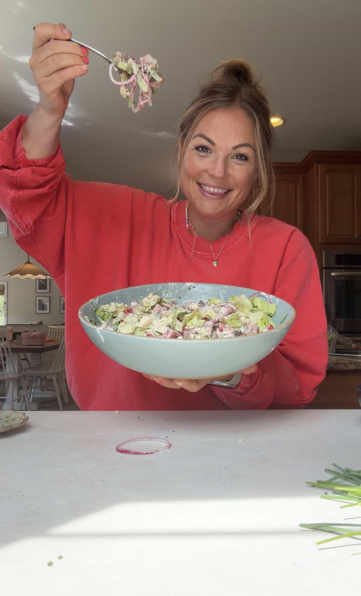 A smiling woman in a red shirt holds a large bowl of salad with one hand and lifts a forkful of salad with the other in a bright kitchen.