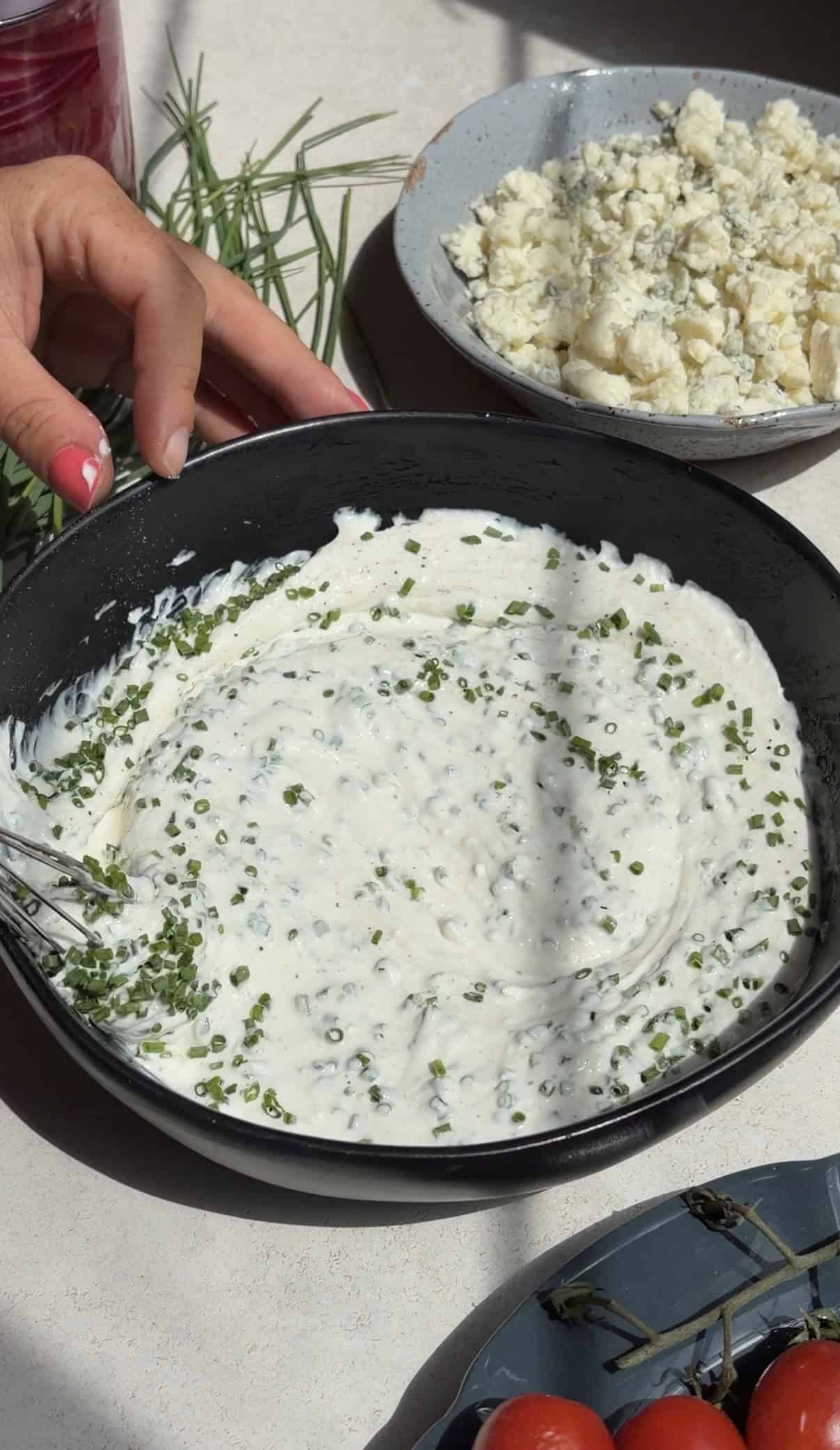 A black bowl filled with creamy blue cheese dressing topped with chopped chives, being mixed with a spoon. A hand with pink nail polish is nearby. In the background, there are plates with cheese and fresh chives.