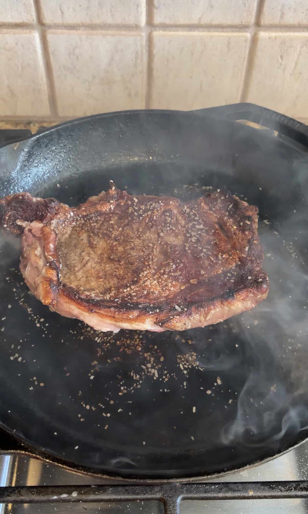 A seasoned steak is searing in a black cast iron skillet, surrounded by smoke, with a tiled backsplash in the background.